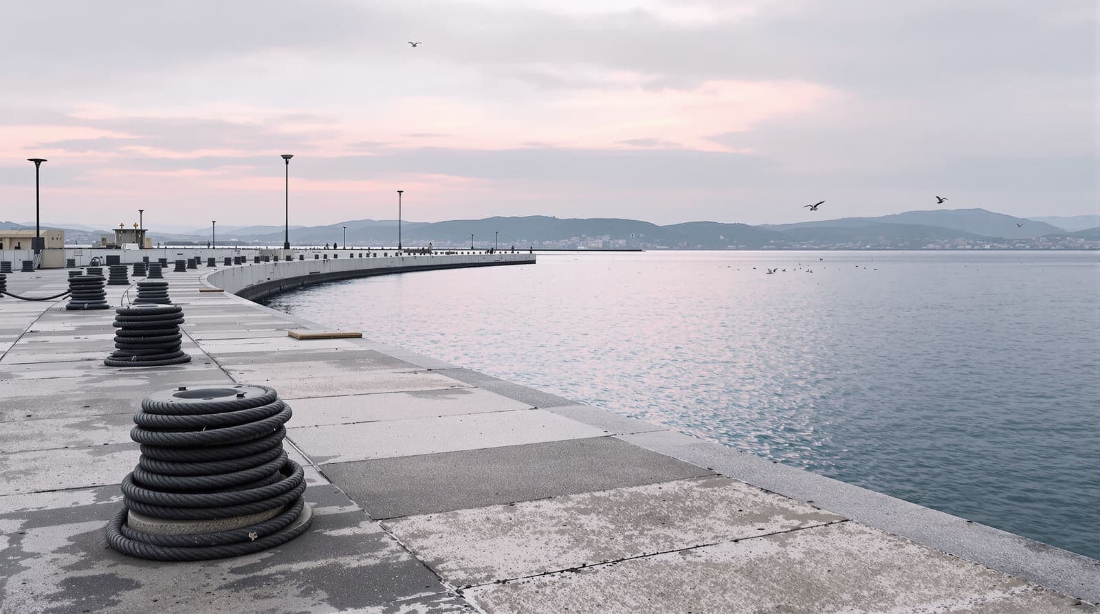 Illustration of an empty naval quay at dawn, mooring bollards and coiled ropes beside calm Mediterranean harbour water.