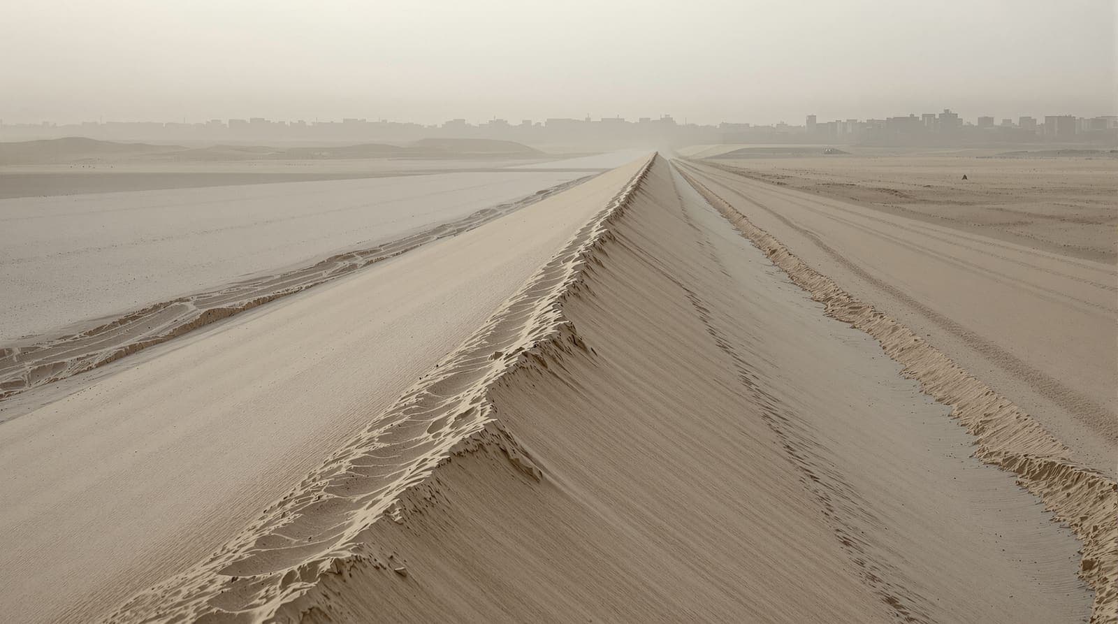 Illustration of a long sand berm and a rutted track across open ground in northern Gaza at dawn, low buildings hazed on the skyline.