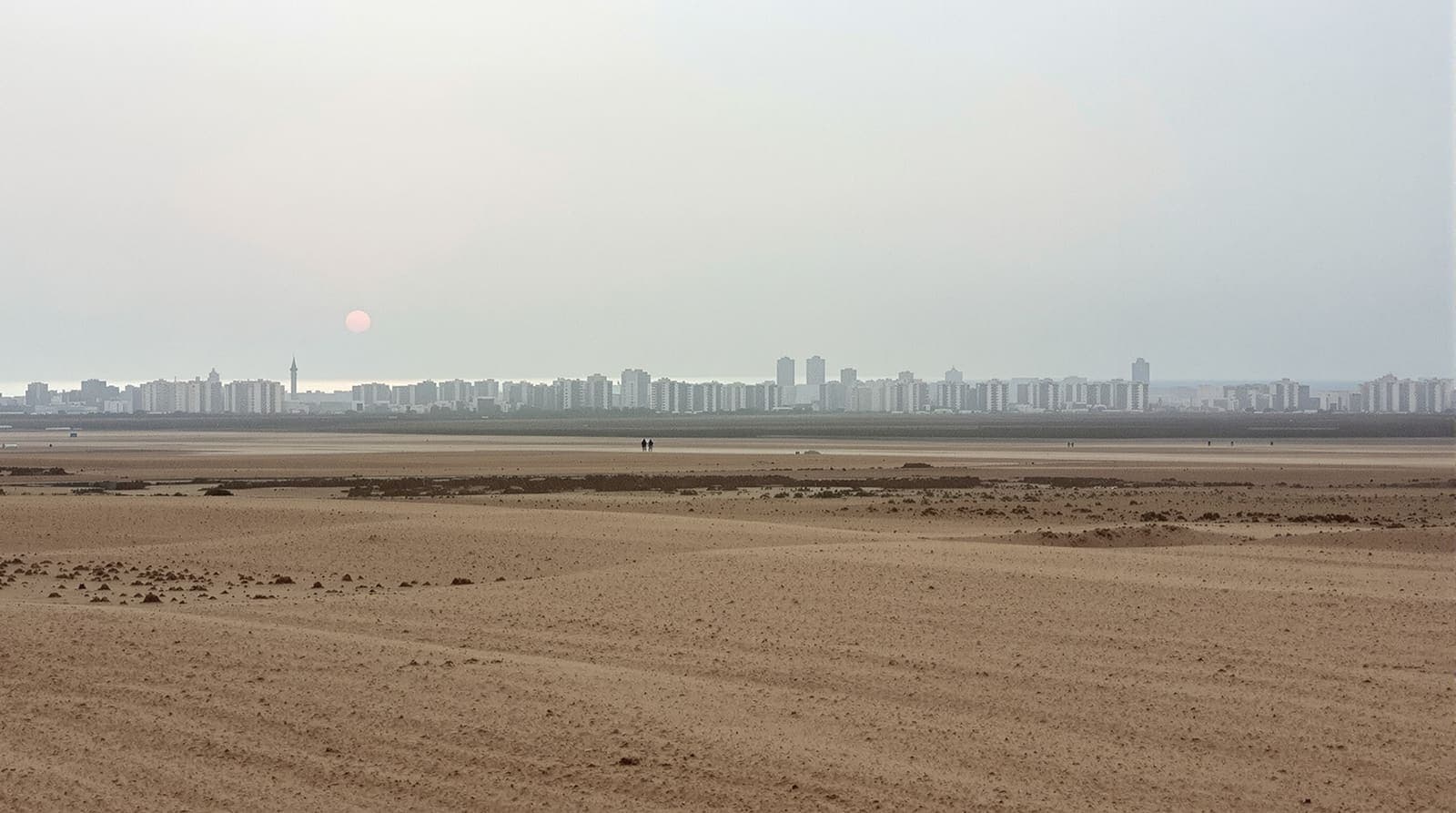 Illustration of the Gaza City skyline in hazy dawn light, seen from a distance across open sandy ground near the coast.