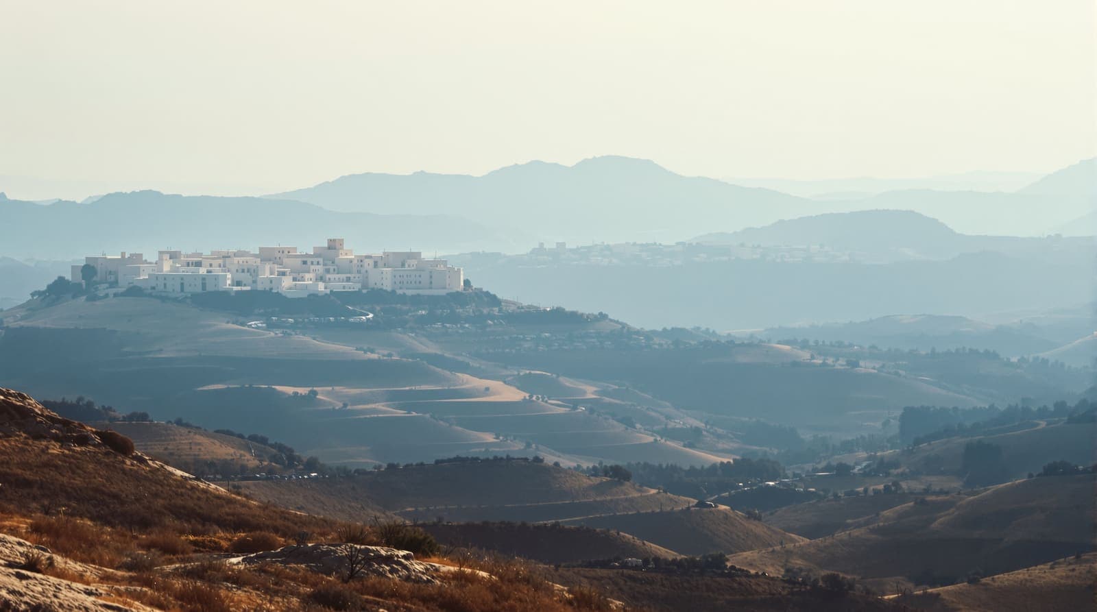 Illustration of a distant hill town in southern Lebanon seen across a hazy valley of olive terraces at first light.