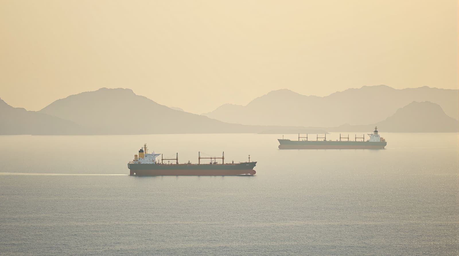 Illustration of oil tankers crossing the calm waters of the Strait of Hormuz at hazy dawn, with mountains in the mist.