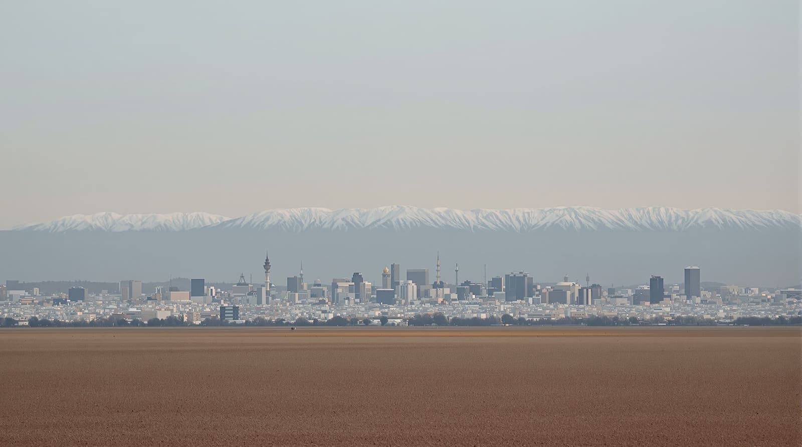 Illustration of a distant city skyline beneath a long mountain ridge, hazy and quiet at dusk.