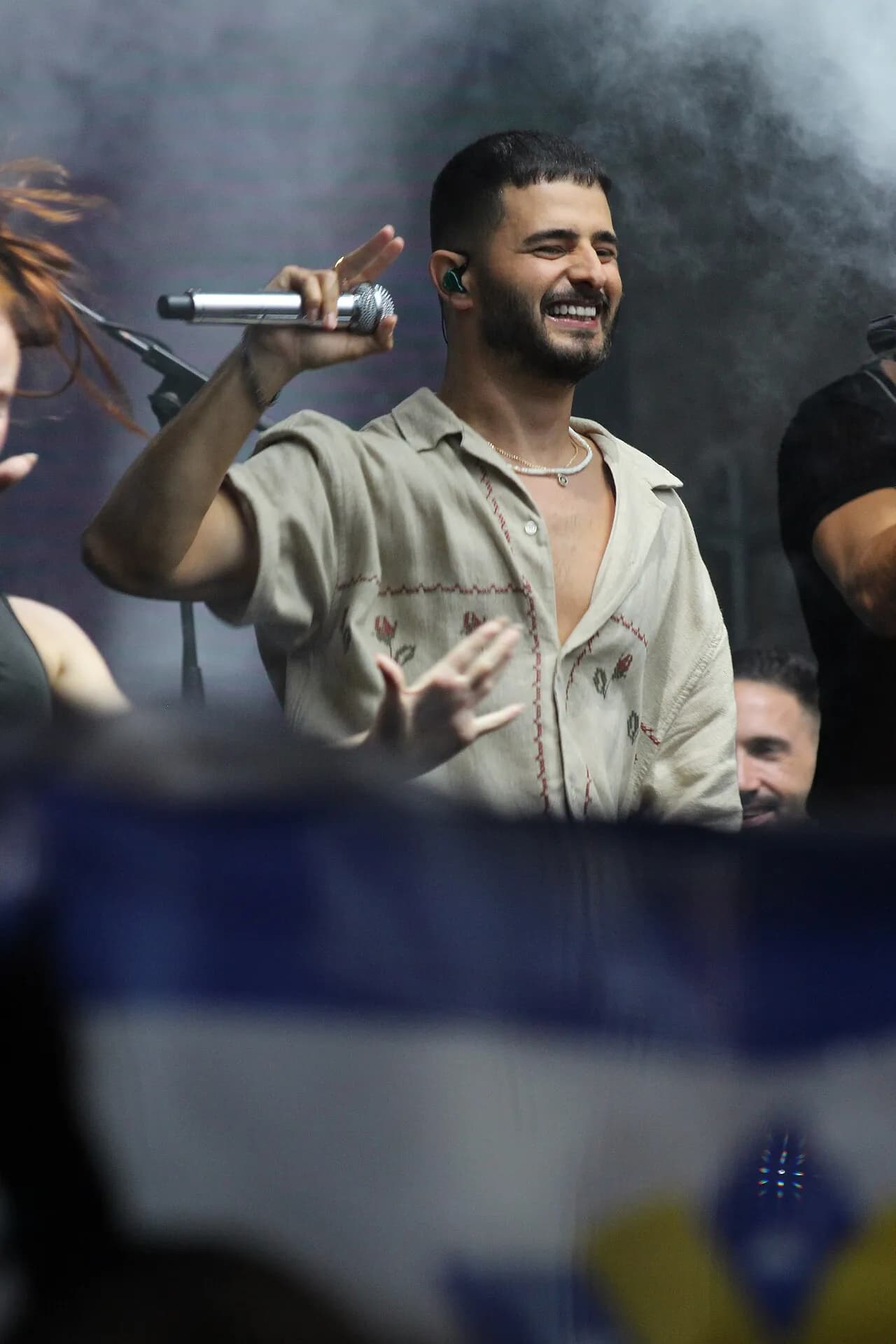 עדן חסון מחייך על הבמה עם מיקרופון, ודגל מטושטש מול הבמה, אוגוסט 2024