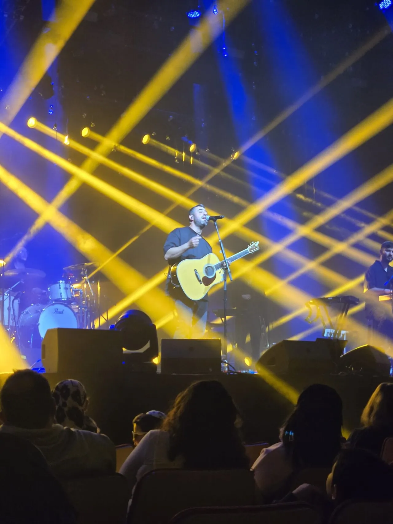 Ishay Ribo with an acoustic guitar under yellow and blue stage lights at Binyanei HaUma, Jerusalem, 2025