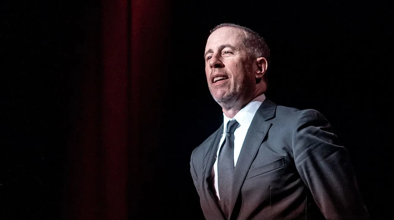 Jerry Seinfeld mid-set on a London stage, in front of a red curtain, 2019