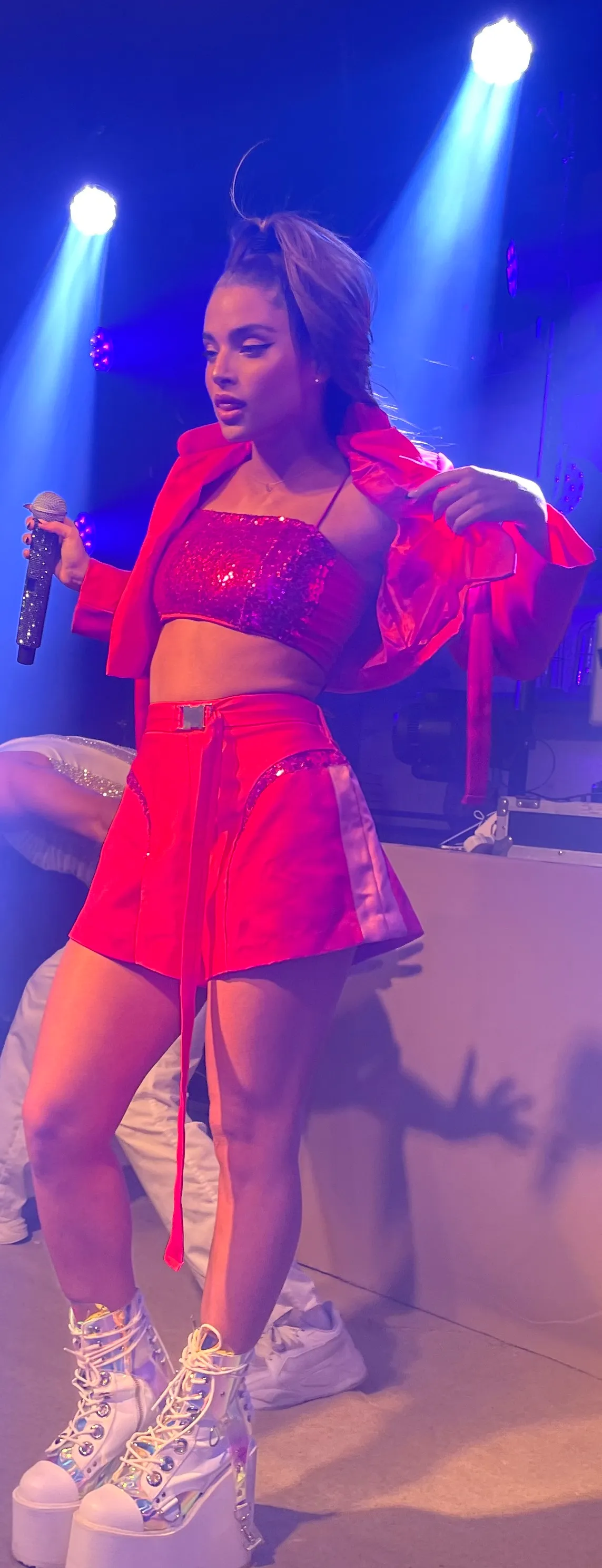 Noa Kirel on stage in a red outfit, holding a microphone under blue spotlights, 2022
