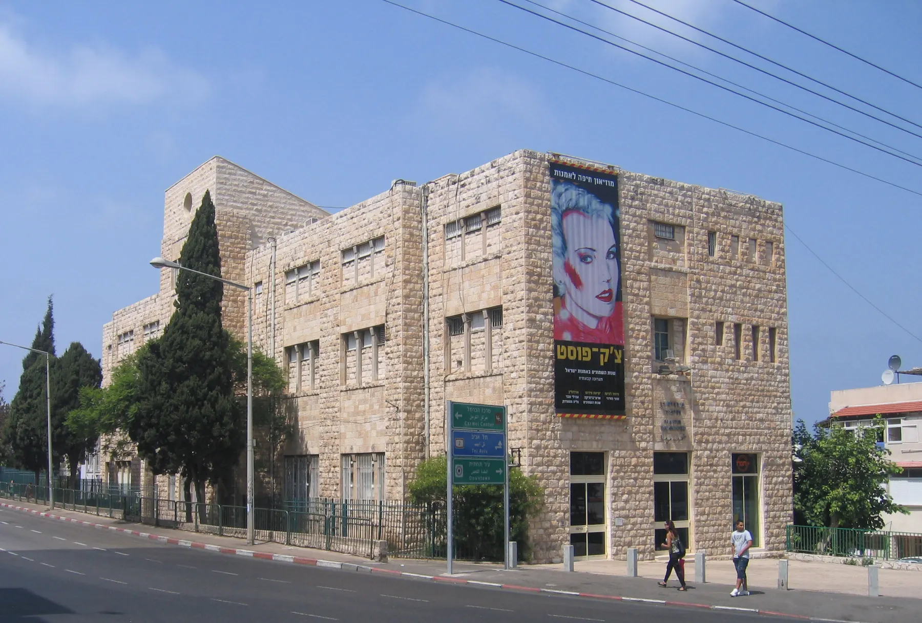 The stone building of the Haifa Museum of Art on a street corner, an exhibition banner on its wall