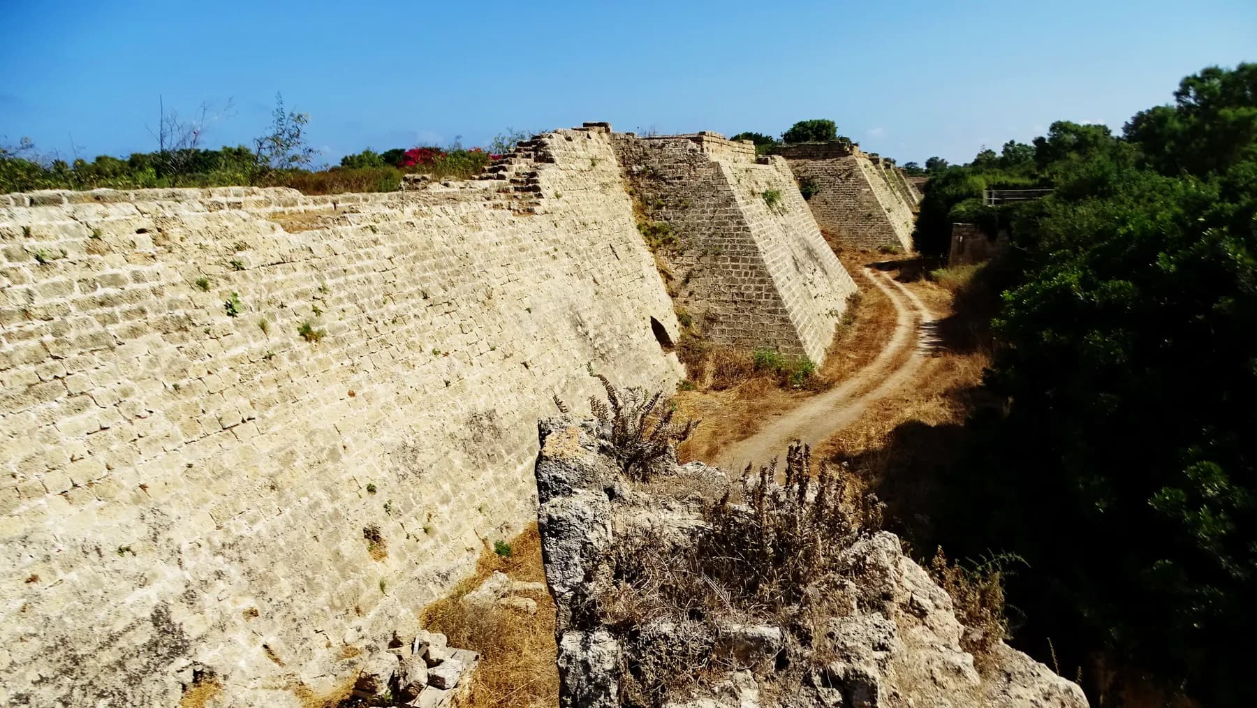 The sloping stone ramparts of the Crusader city at Caesarea rising on both sides of a dry moat, with a dirt track running along its floor