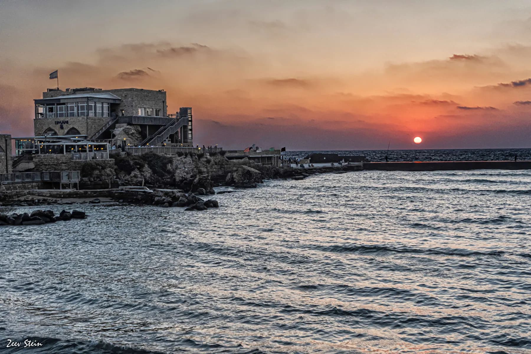 Sunset over the harbour at Caesarea, with a low stone breakwater reaching out into the Mediterranean and the citadel headland on the left