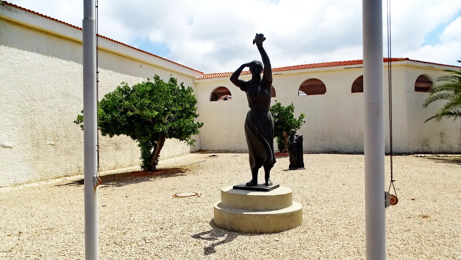A dark bronze figure with one arm raised, standing on a round stone plinth in a gravel courtyard enclosed by a white arcaded wing with a red-tiled roof