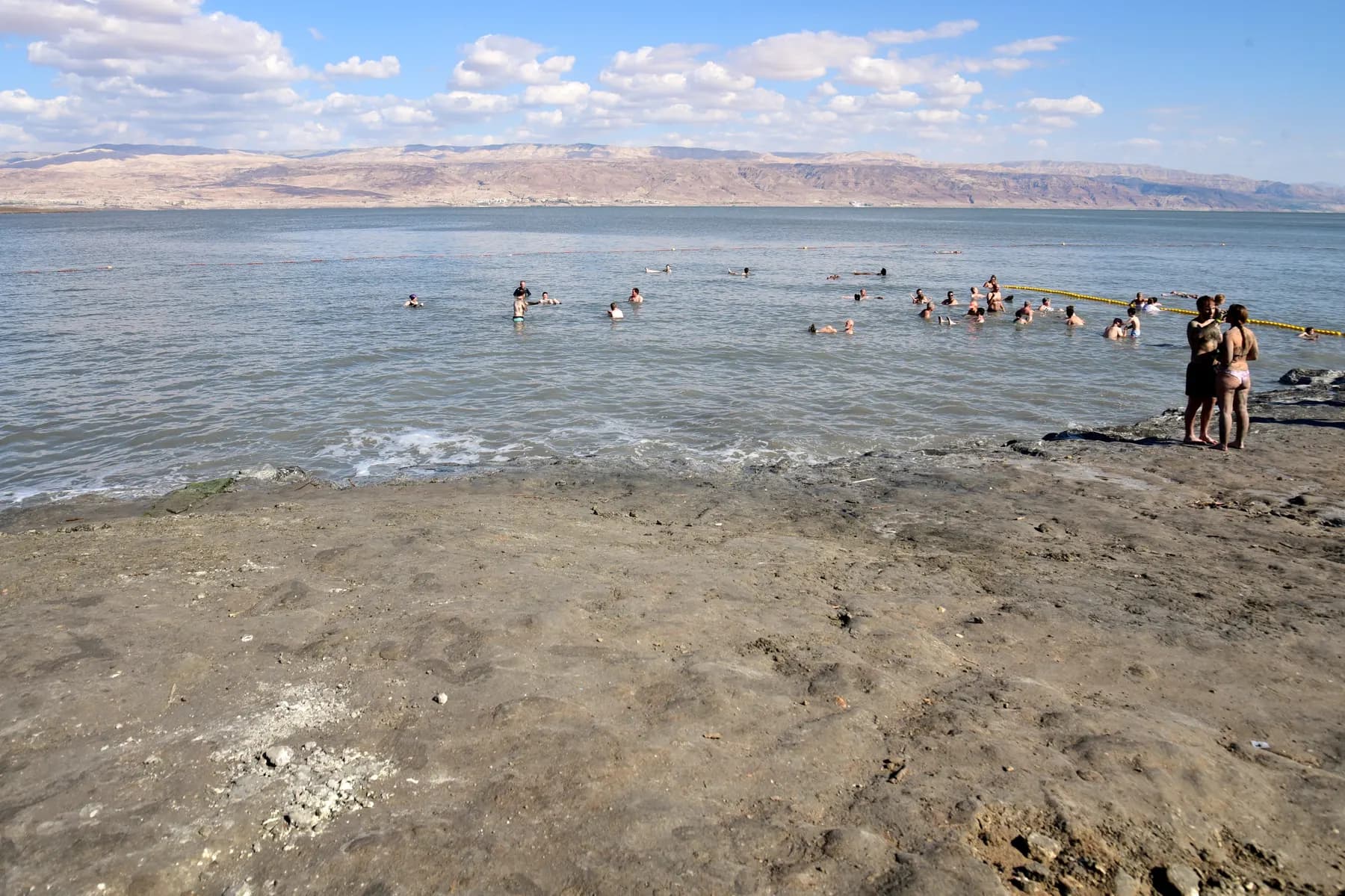 A broad grey mud and salt shore at the Dead Sea with bathers floating in the still water beyond and a bare mountain ridge on the far side