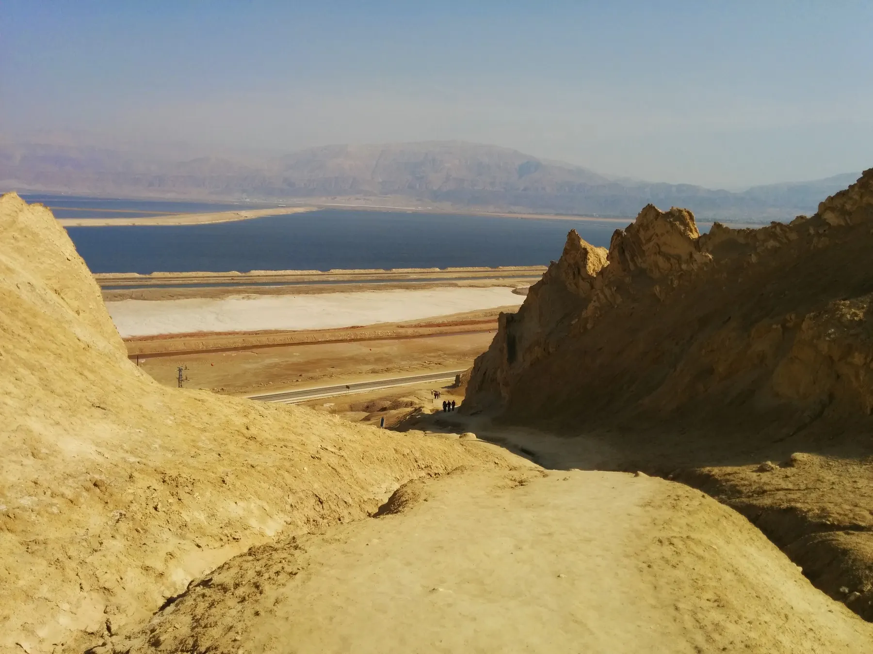 Eroded pale rock slopes of Mount Sodom falling to the Dead Sea, with a road and salt flats along the shore and hazy mountains beyond the water