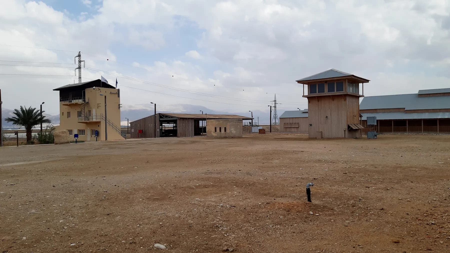 A timber lookout tower and low stone and timber buildings standing around a wide empty gravel yard beneath a cloudy sky