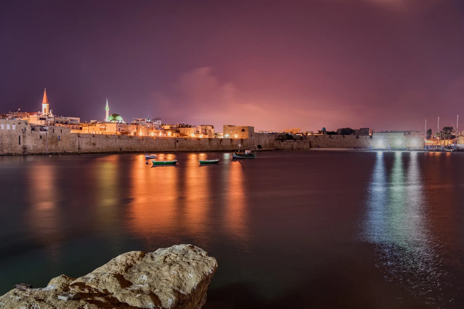 The lit stone sea walls and minarets of Akko's old city seen at night across the harbour, with small boats moored on the water