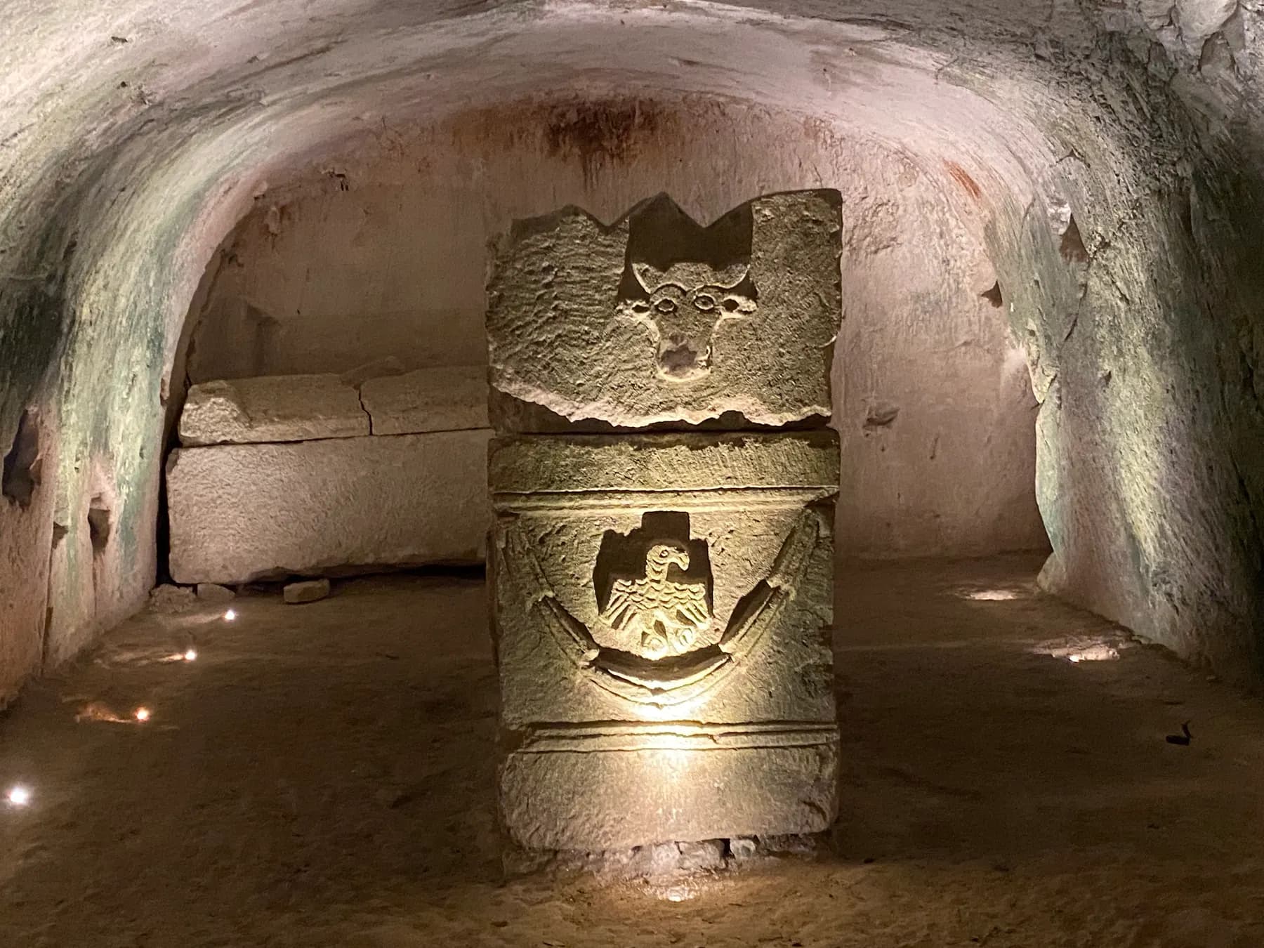 A carved stone bearing a bull's head above an eagle with garlands, lit inside a vaulted rock-cut burial chamber, with a plain sarcophagus against the wall behind