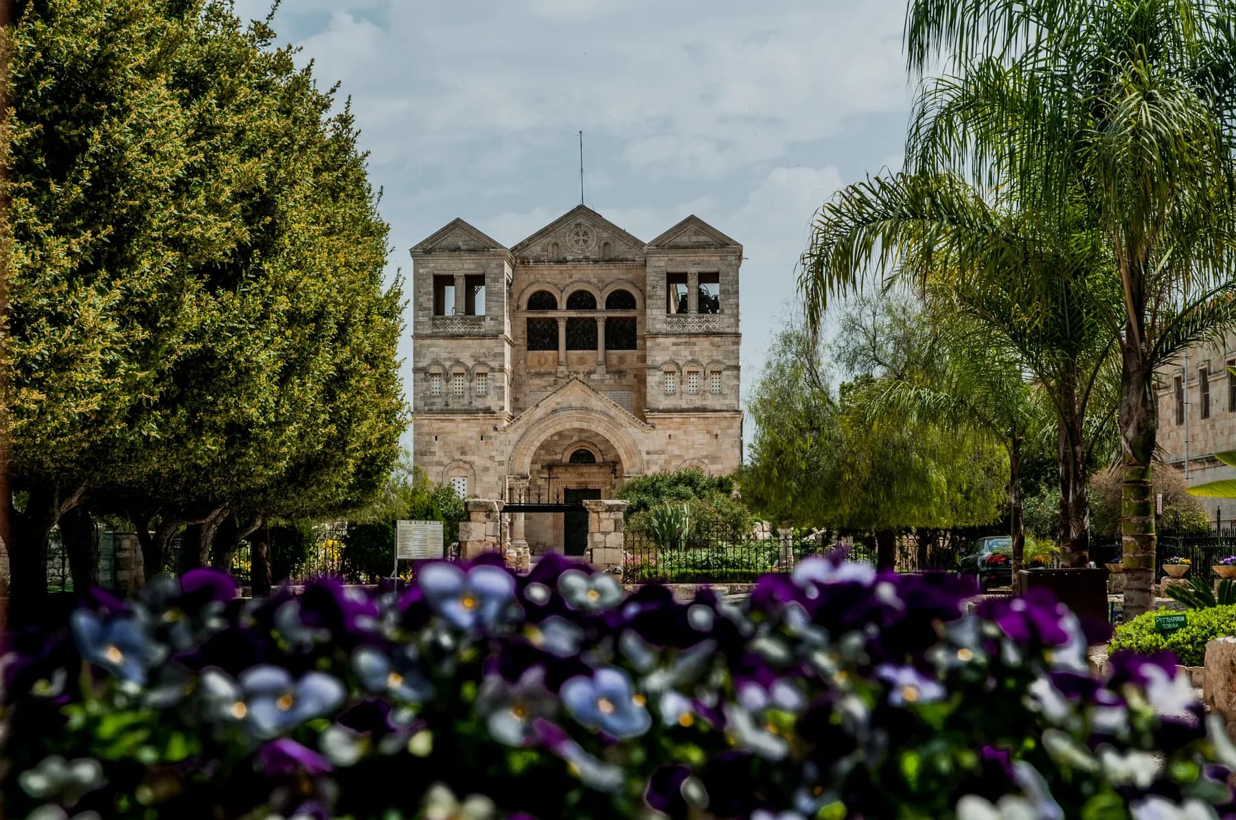 The stone facade and twin towers of the Basilica of the Transfiguration on Mount Tabor, seen across a garden bed of purple pansies