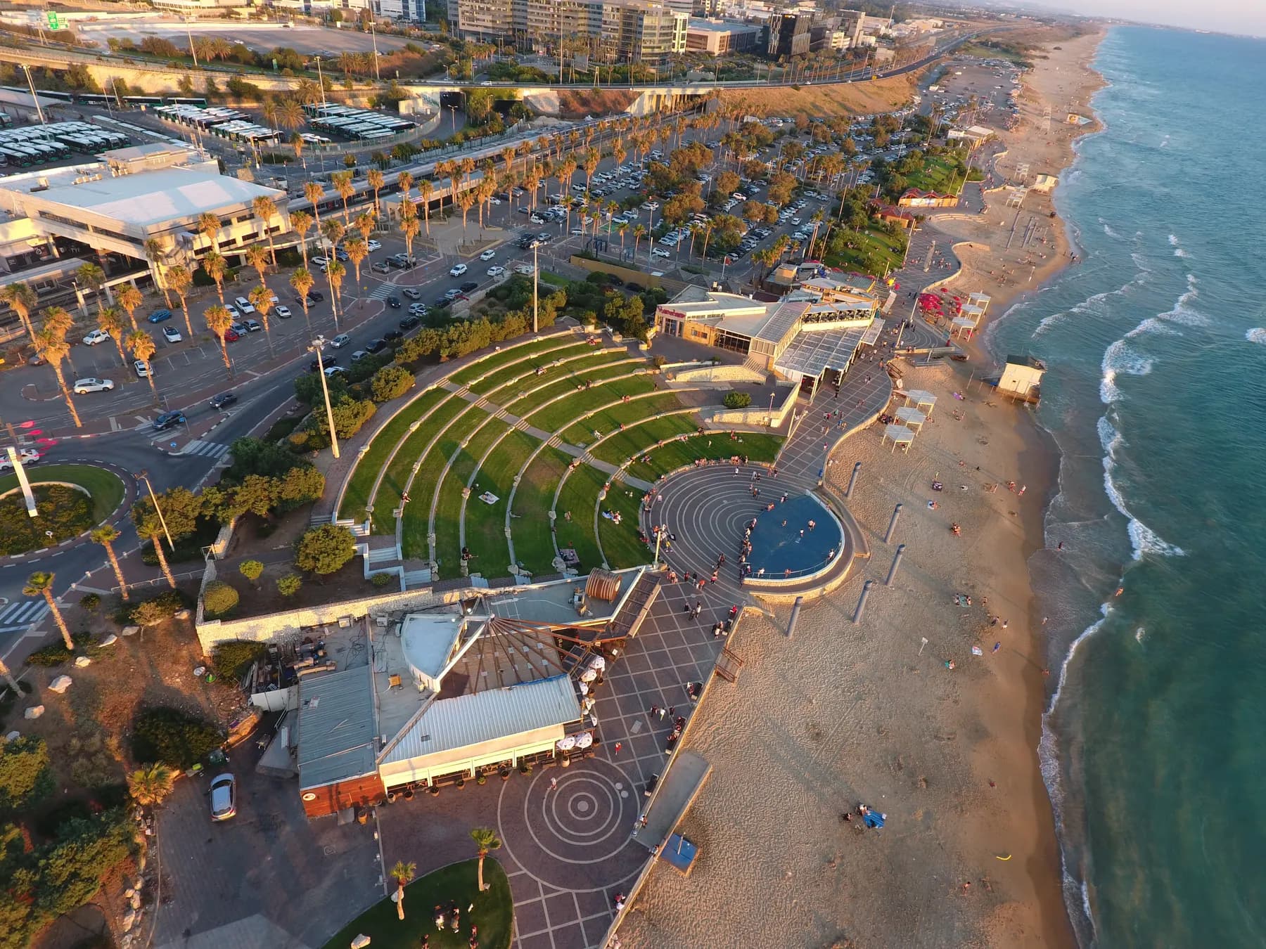 Golden-hour aerial of the Dado Beach promenade in Haifa, with grass amphitheatre terraces and a circular paved plaza beside the sand and breaking surf