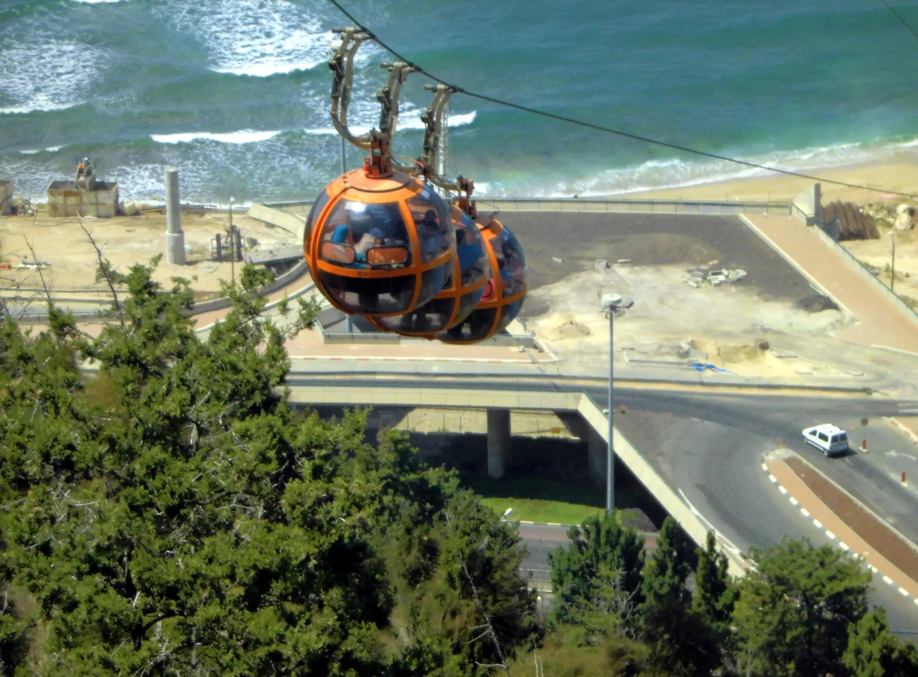 Two orange spherical cable car gondolas hanging from the cable above a coast road and flyover, with the Mediterranean surf breaking beyond