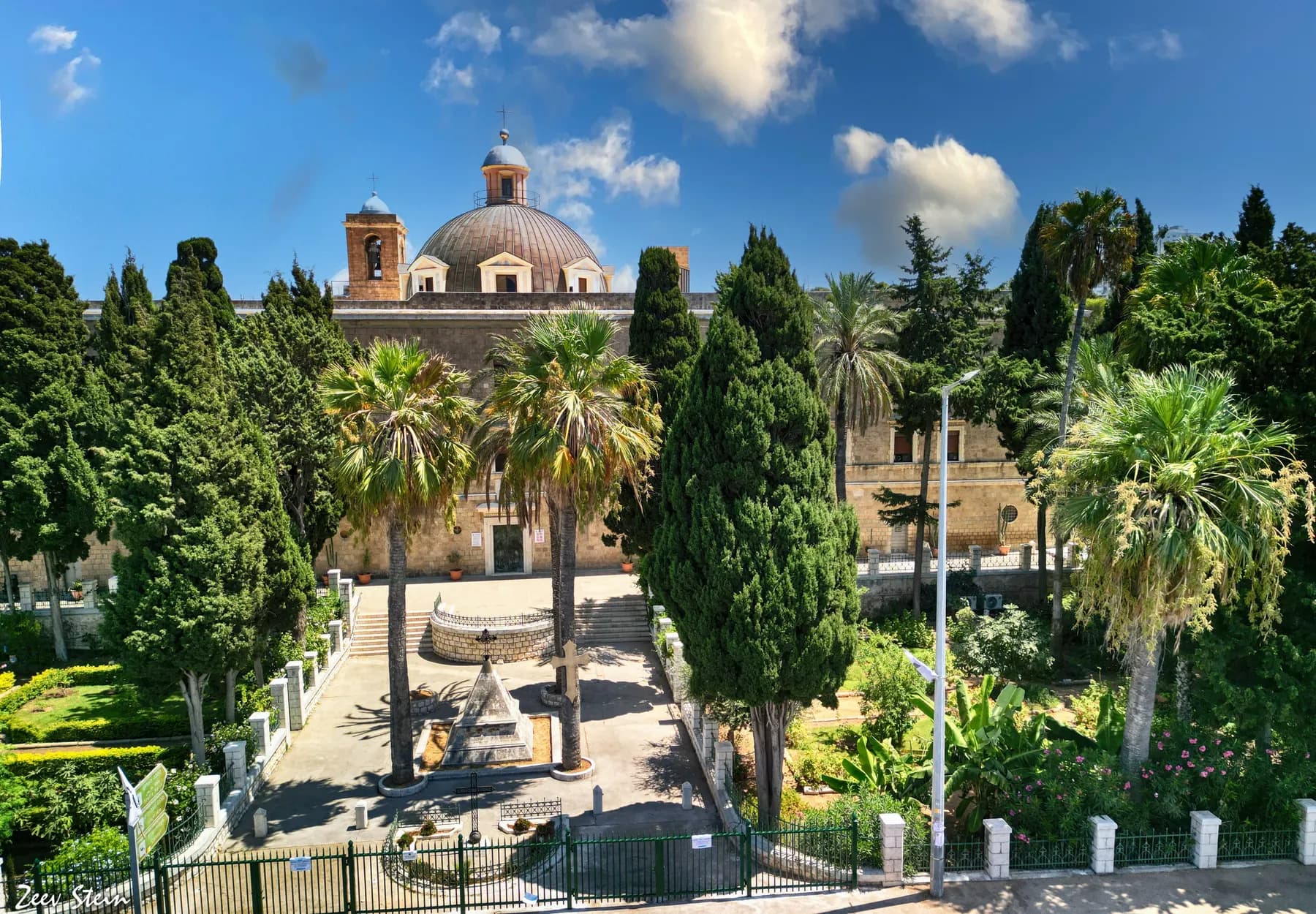 The domed church and bell tower of the Stella Maris monastery on Mount Carmel, seen across its walled garden of palms and cypresses under a blue sky