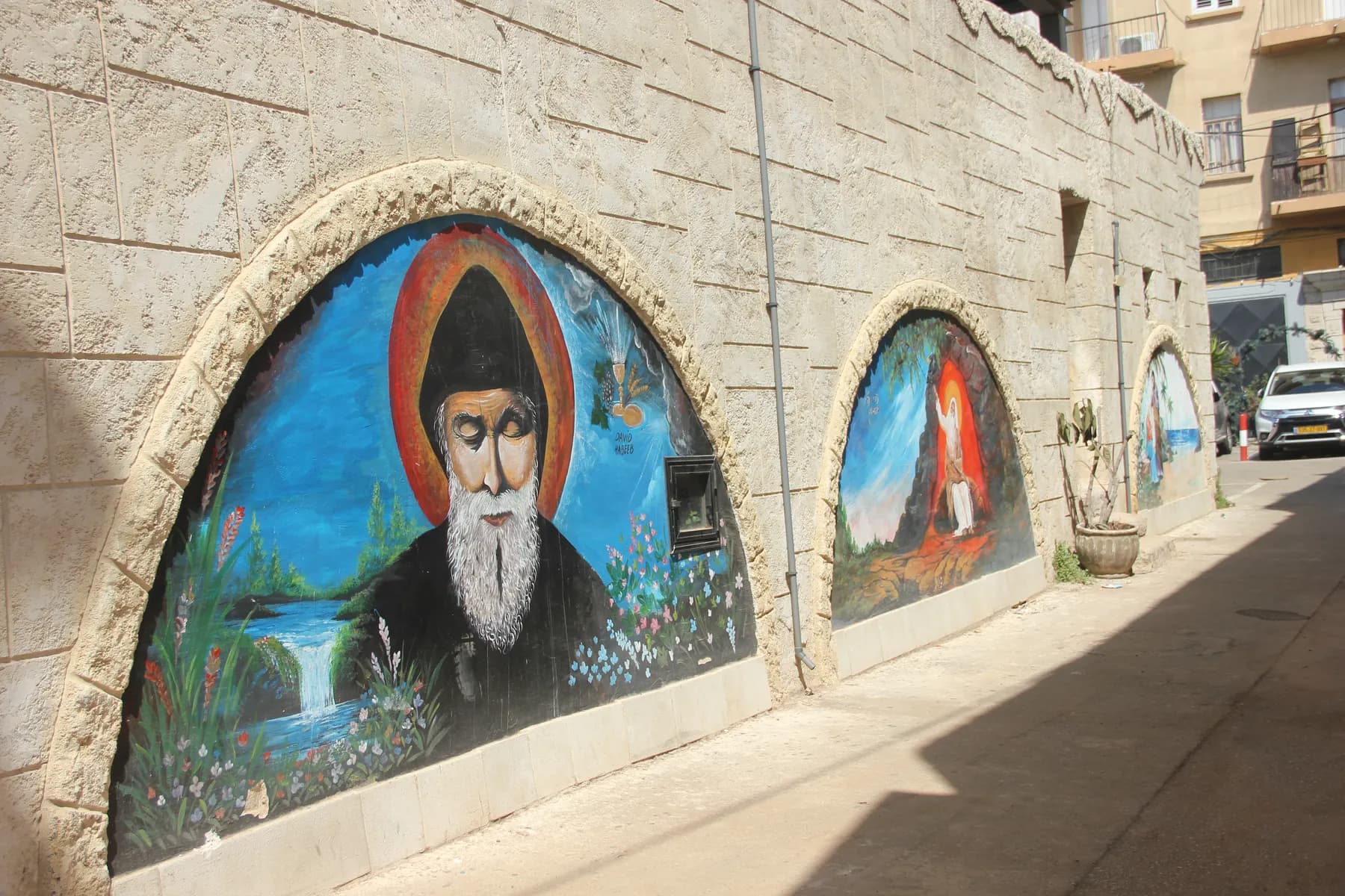 Painted murals set into arched recesses along a rendered stone wall in a narrow Haifa alley, the largest showing a bearded monk in black robes against a blue landscape