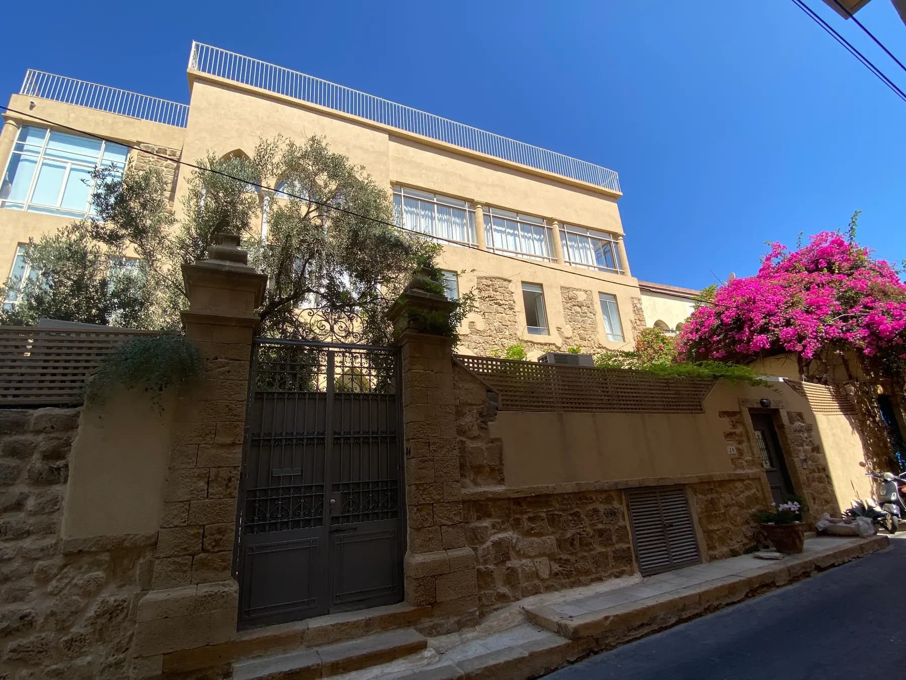 A restored sandstone house behind a high stone wall and dark iron gate on a narrow Jaffa lane, with bougainvillea flowering over the wall to the right