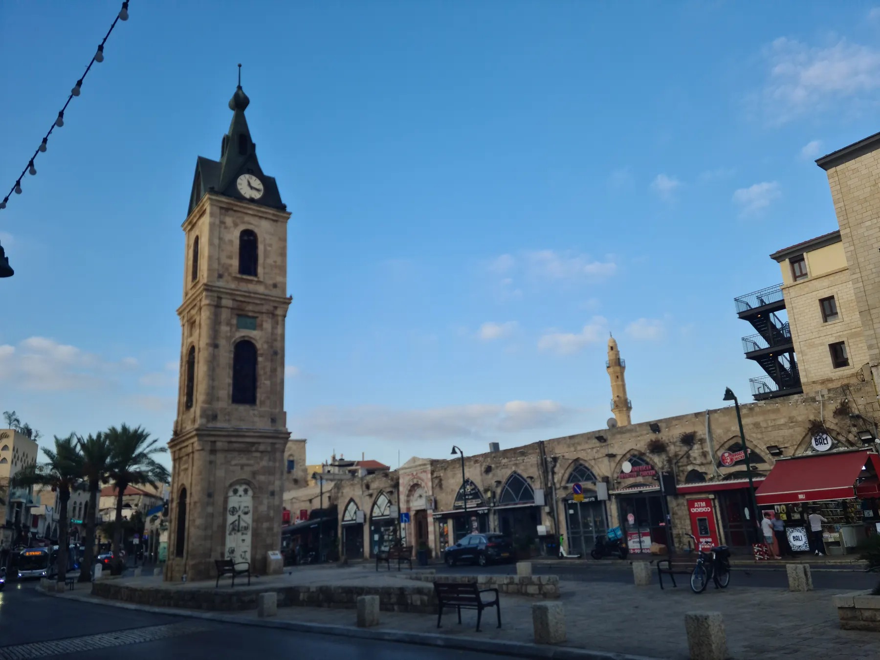 The Ottoman stone clock tower with its dark spire standing in Jaffa's empty square in early morning light, with palm trees and arcaded stone buildings behind it