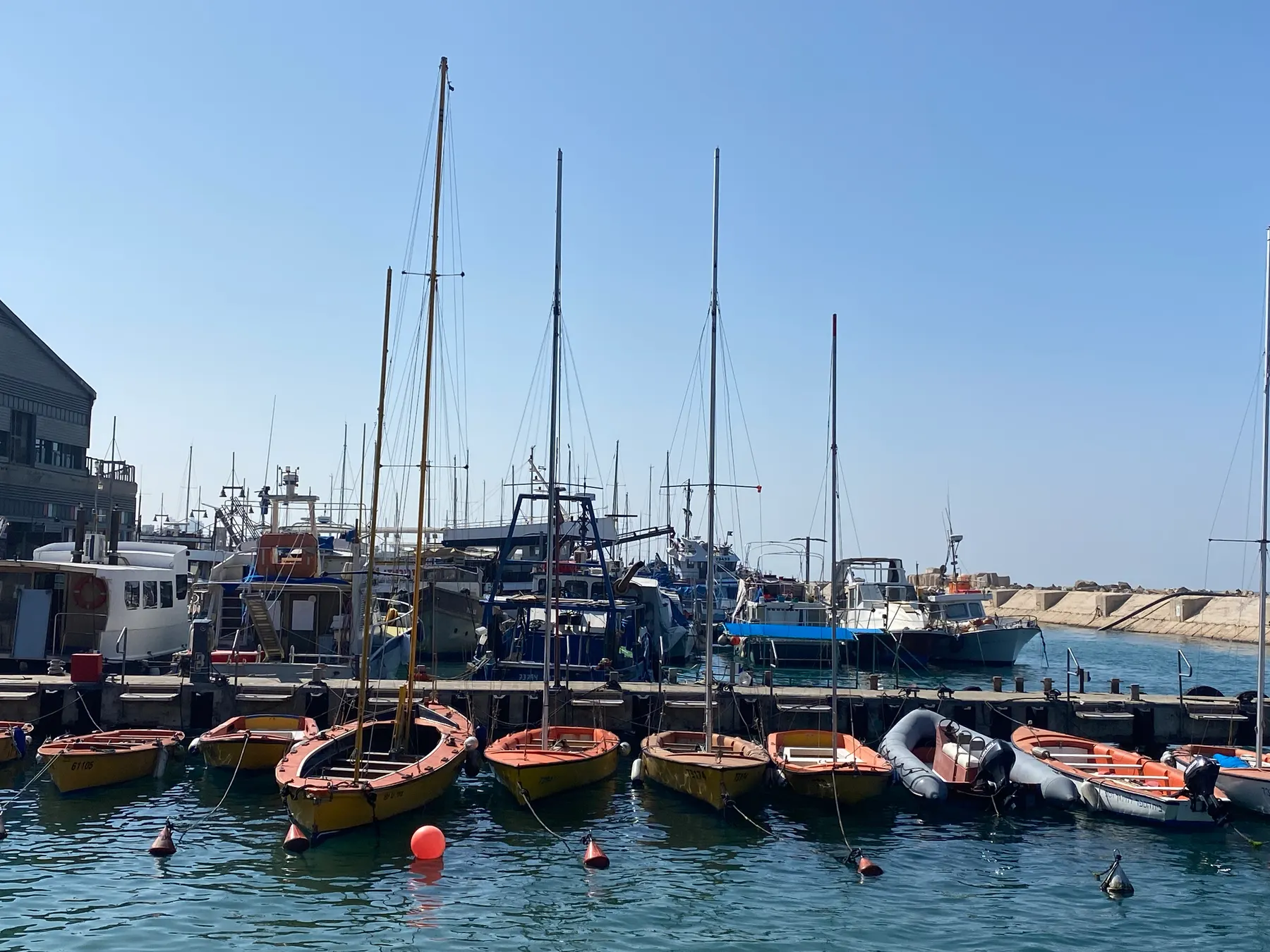 Fishing boats and tall masts moored along the quay at Jaffa harbour, with small orange dinghies on the water and the stone breakwater running out to the sea beyond