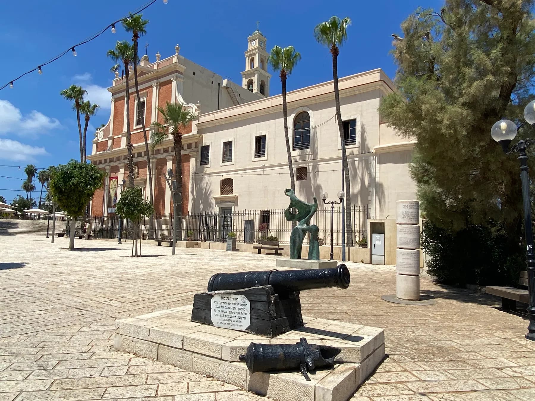 The red brick facade and pale bell tower of St Peter's Church in Jaffa seen across an empty paved square, with palm trees in front and an old cannon mounted on a stone plinth