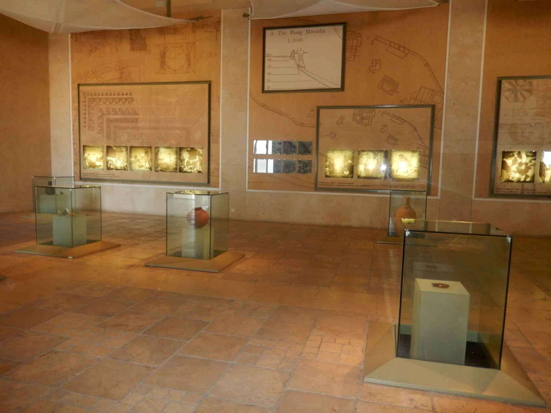 Freestanding glass cases holding pottery in a museum gallery, with lit wall niches of small finds and excavation plans of Masada drawn on the stone walls