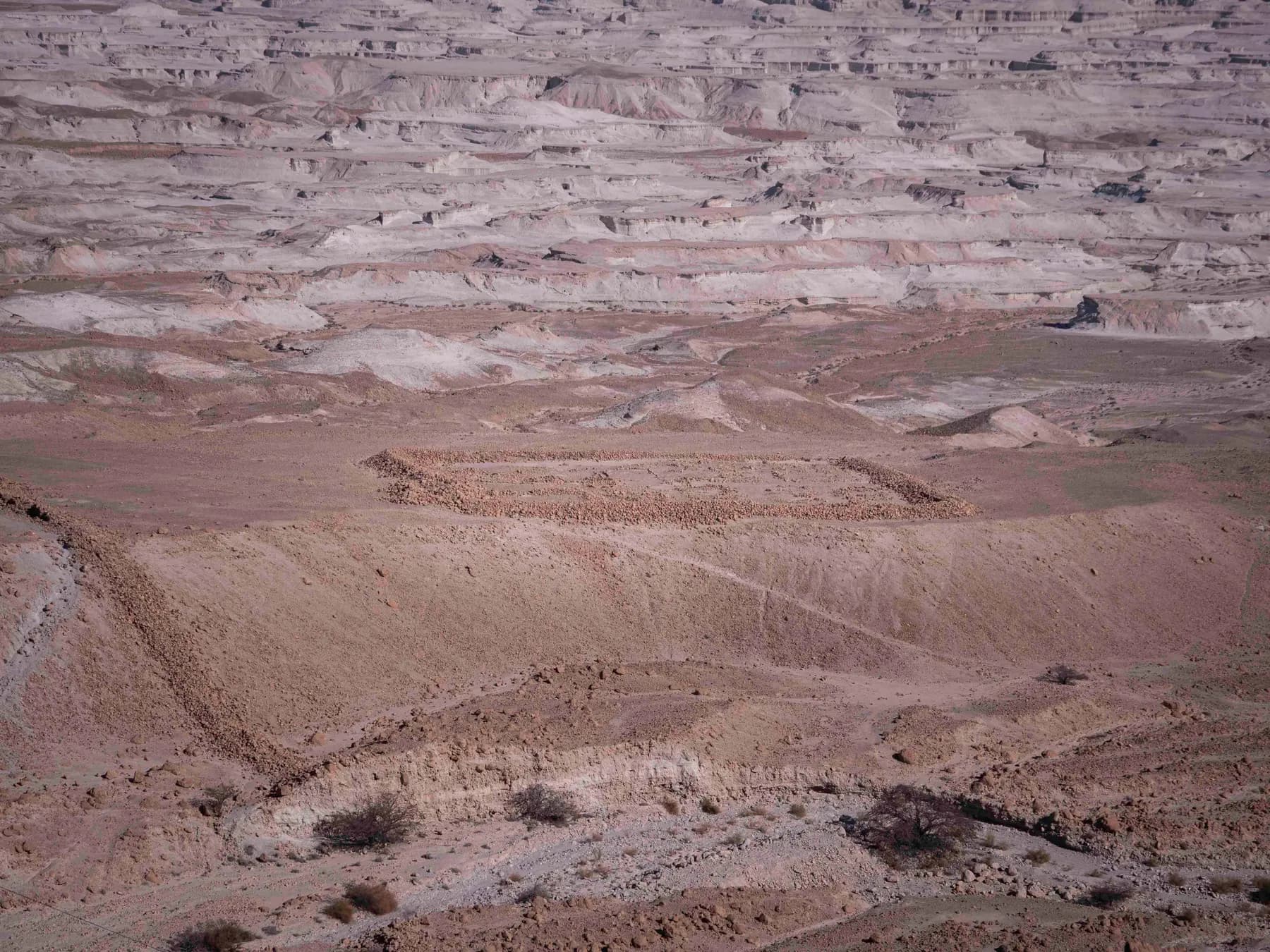 The rectangular stone rampart of a Roman camp laid out on a desert terrace below Masada, with pale eroded badlands stretching behind it