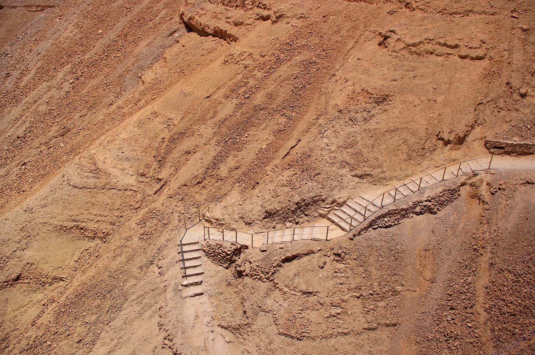 A hairpin bend of a railed footpath cut into the bare red rock of the Masada cliff face, switchbacking up the slope