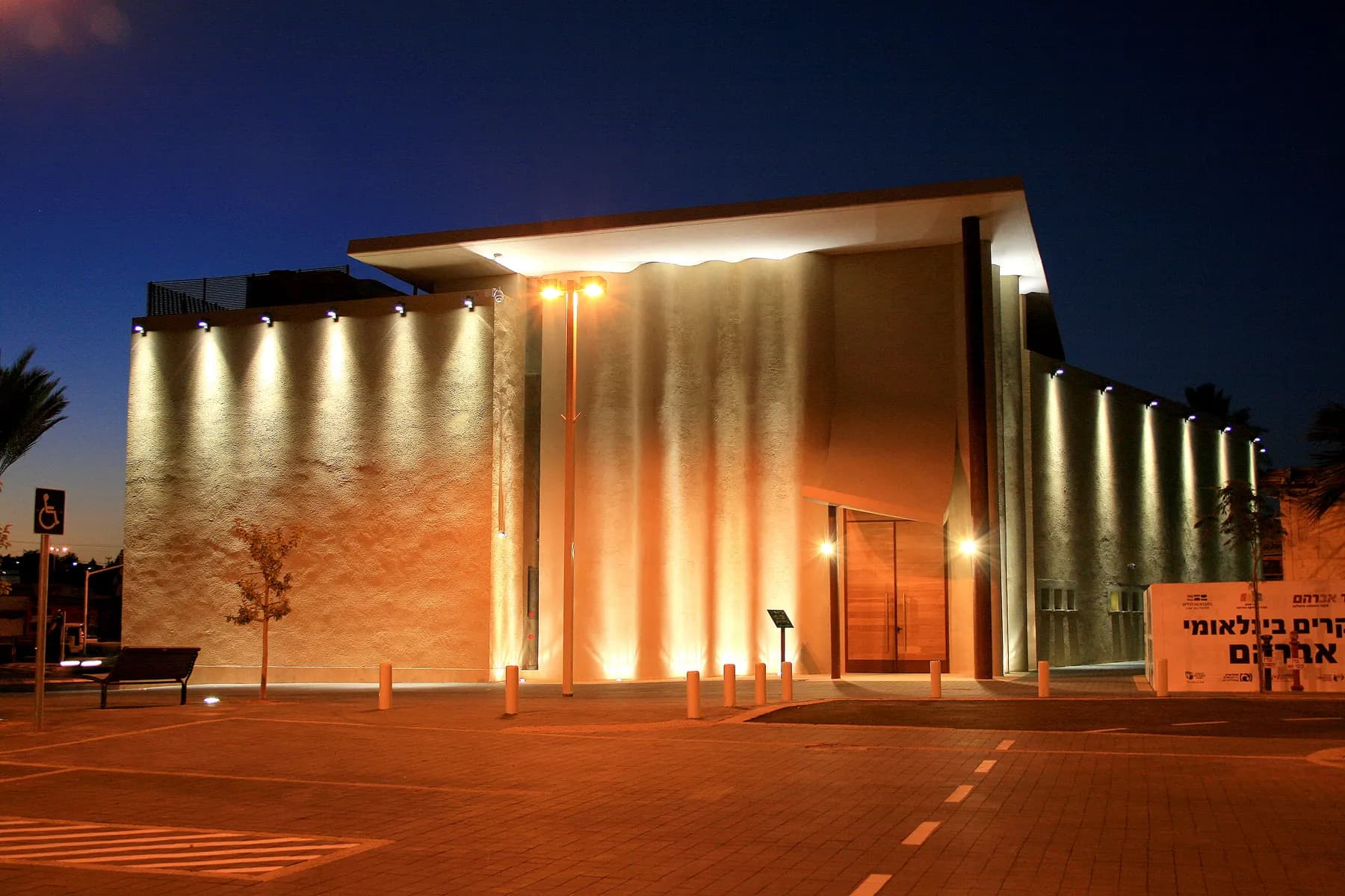 The fluted concrete facade of the Abraham's Well visitor centre in Beersheba lit by uplighters at dusk, above an empty paved plaza