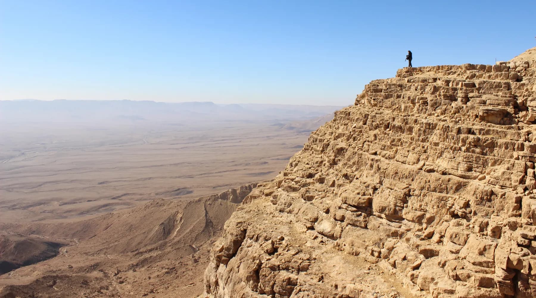 The layered limestone cliff of the crater rim at Makhtesh Ramon dropping away to the pale desert floor below, with a lone figure standing on the clifftop