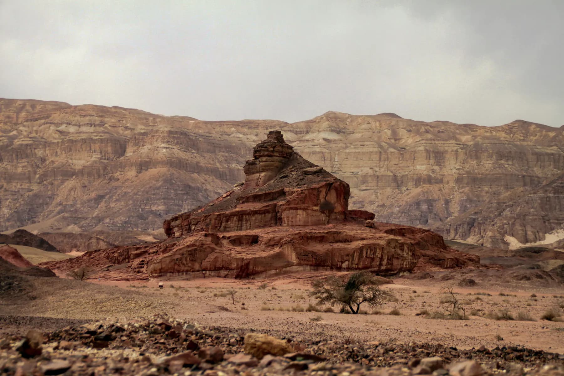A stepped red sandstone hill rising from the desert floor at Timna, with banded tan and grey cliffs running across the background