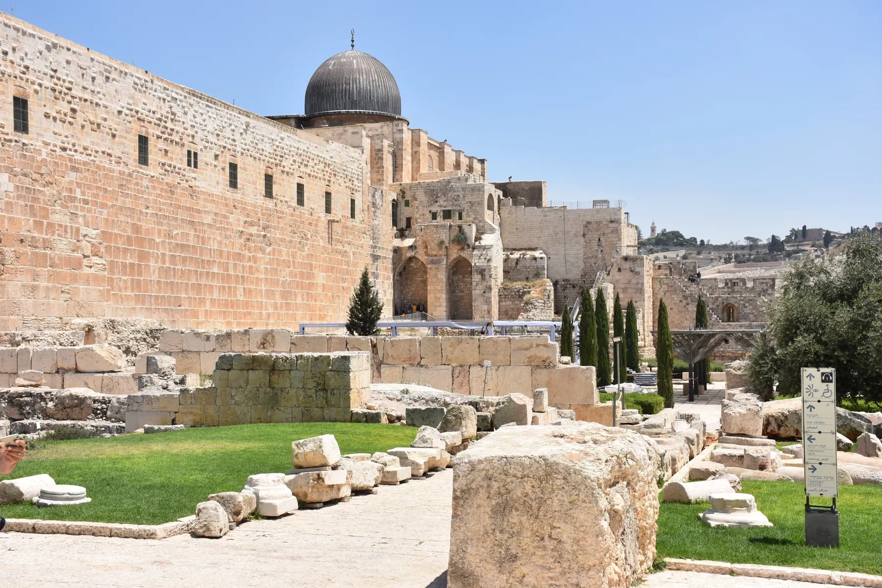 Excavated stone walls and fallen blocks on the grass at the foot of the Temple Mount's high southern wall, with the dark dome of Al-Aqsa Mosque above