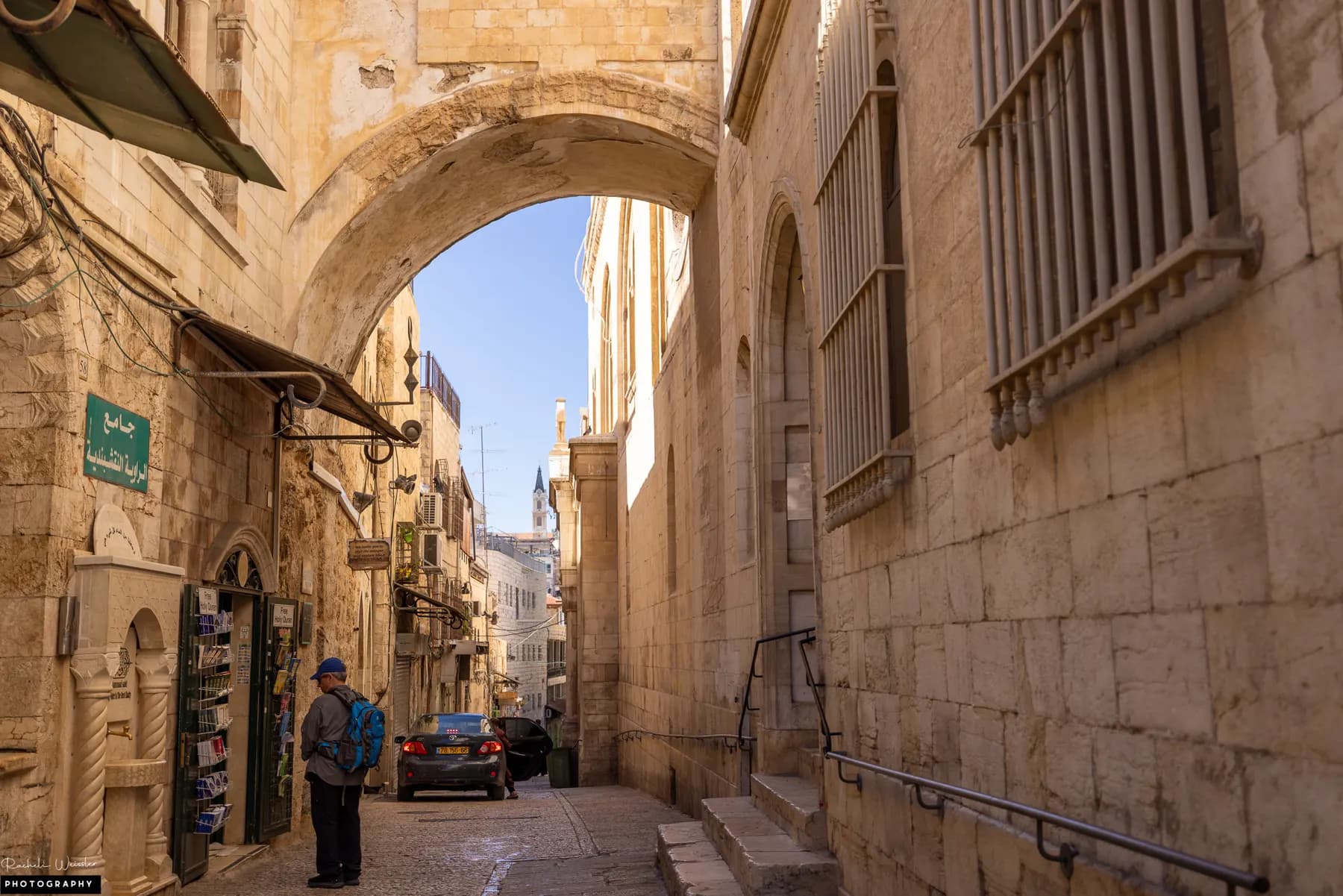 A narrow stone lane in Jerusalem's Old City running beneath a masonry arch, with shop awnings and a lone passer-by along the left wall