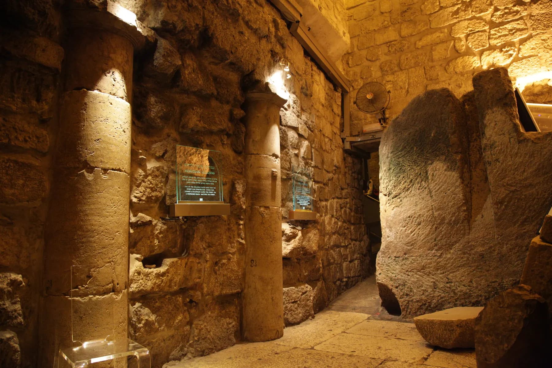 A lamplit underground passage of large stone blocks in Jerusalem, with columns set into the left wall and bare rock rising on the right