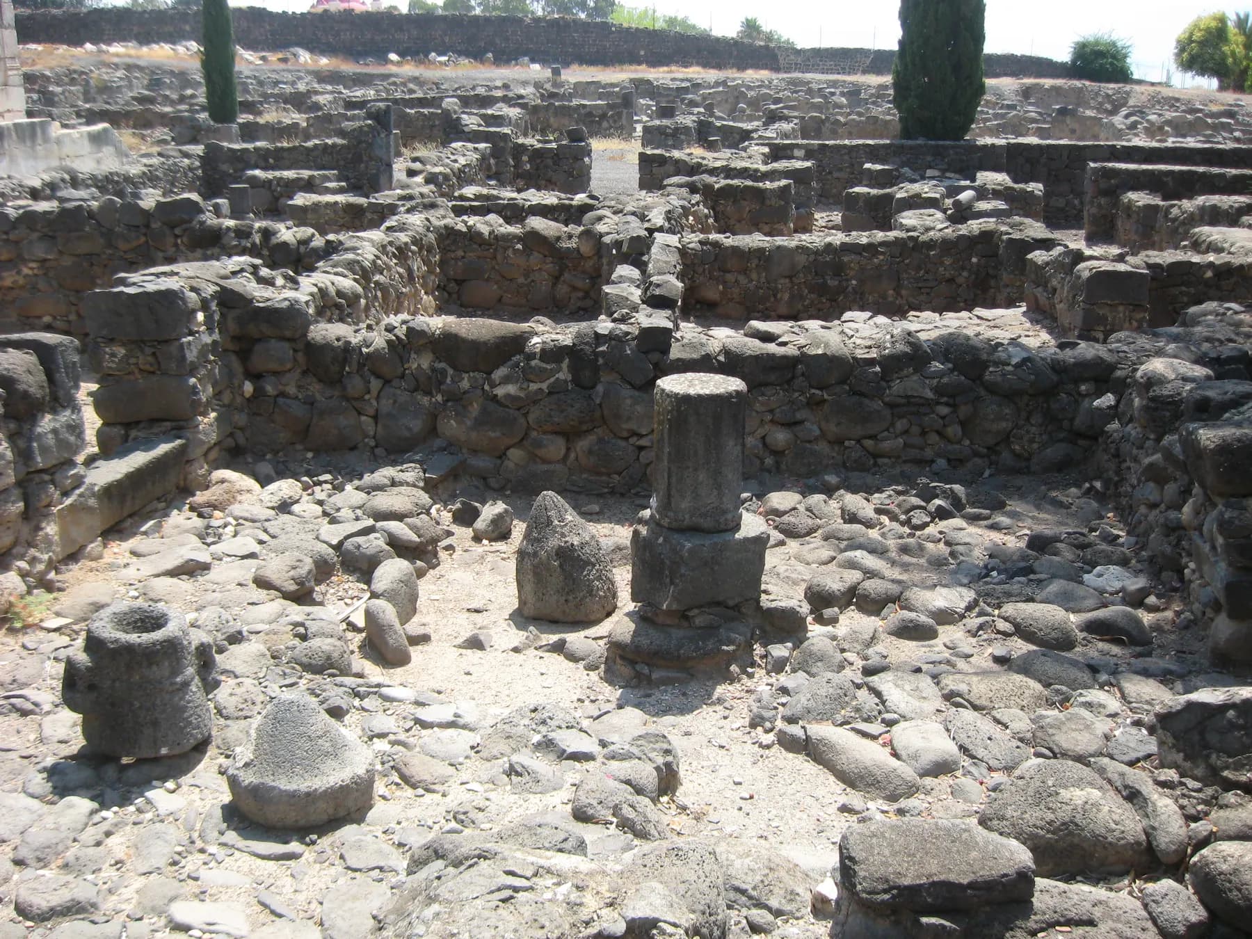 Black basalt walls and scattered millstones in the excavated house ruins at Capernaum, with a column drum standing among them