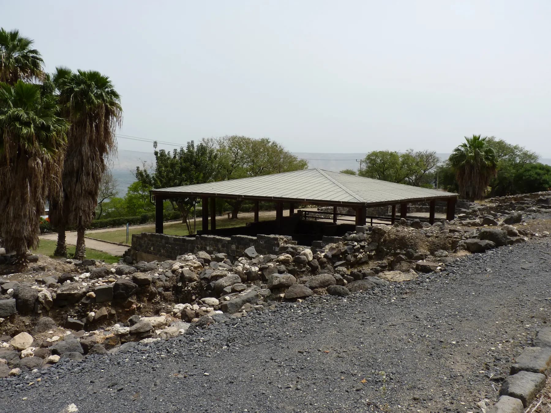 A low metal shelter roof on open posts standing over excavated stone remains at Hamat Tiberias, with rough basalt wall foundations in front and the hazy Sea of Galilee behind