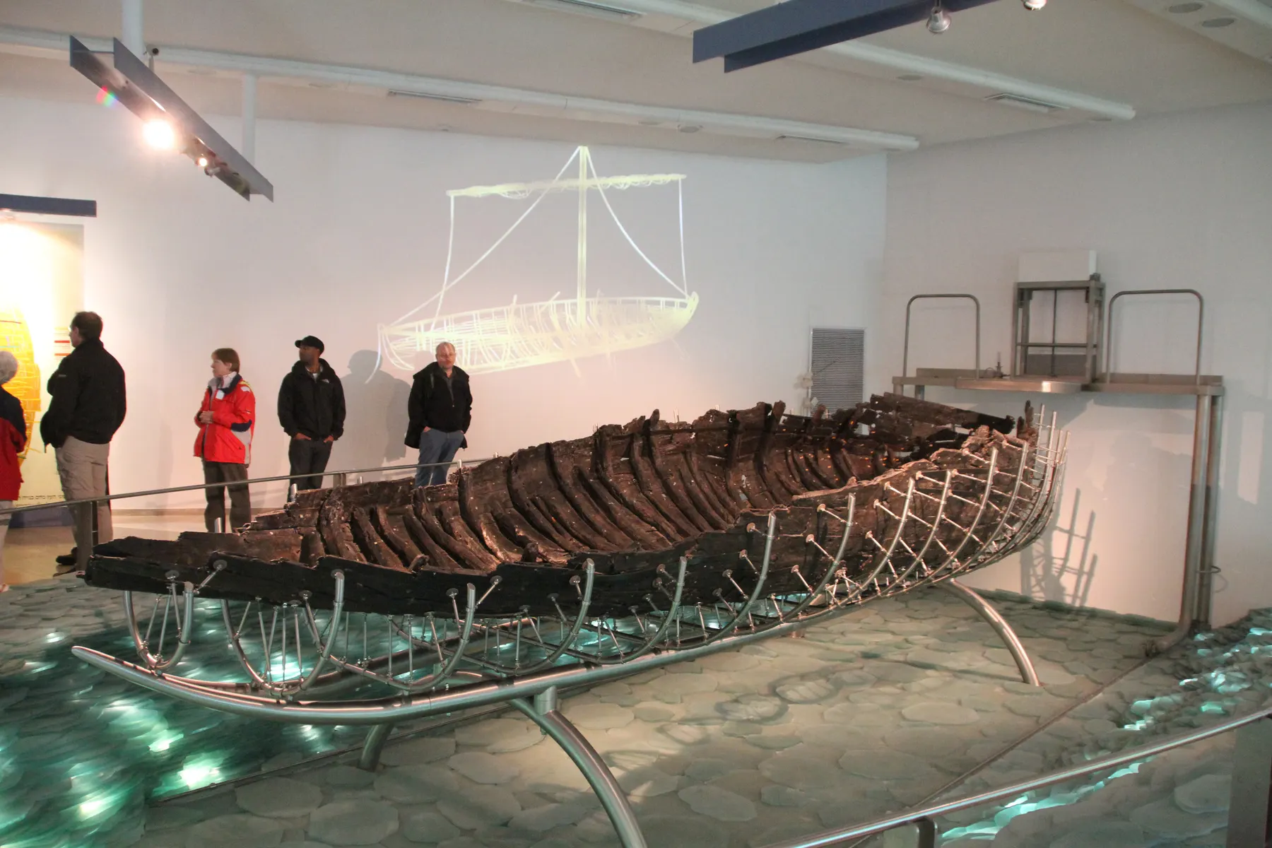 The dark excavated hull of the ancient Galilee boat resting on a steel cradle in a museum hall, with a wire outline of the complete vessel mounted on the wall behind it