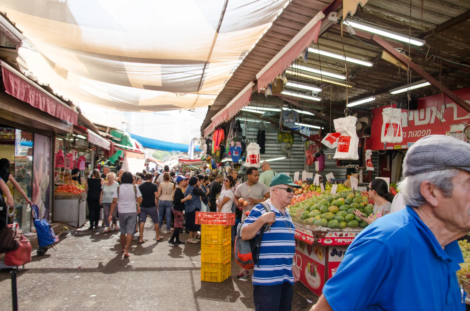 Shoppers filling a market lane between fruit stalls and clothing stands, under white awnings stretched overhead