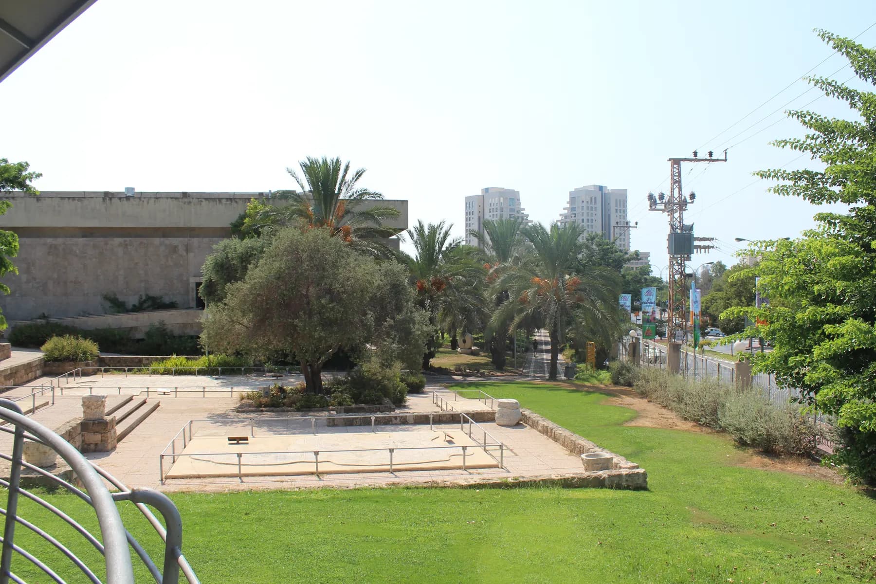 A low stone-faced pavilion of the Eretz Israel Museum above a paved court where stone column drums stand behind metal railings, an olive tree at its centre