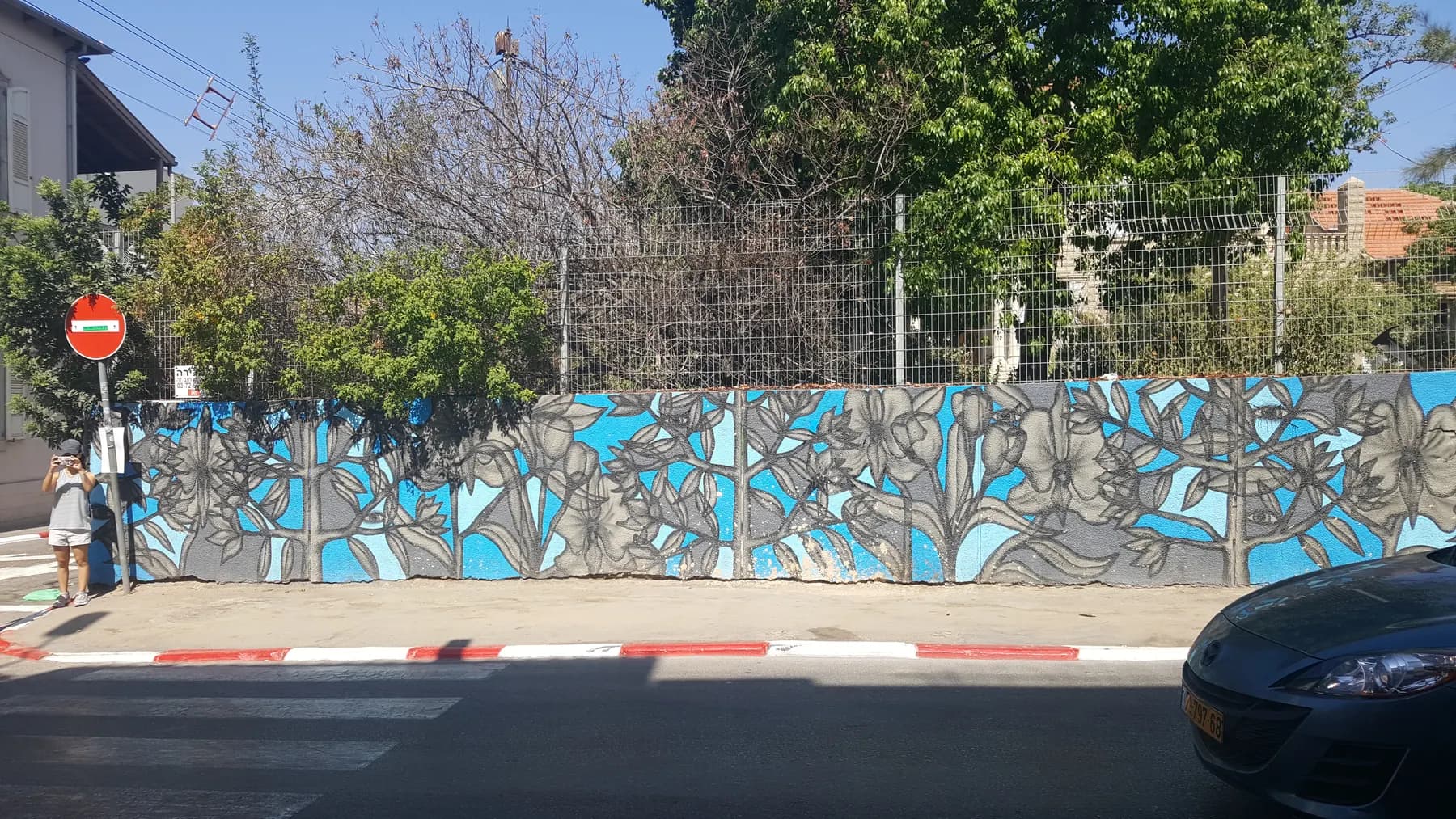 A long street wall in Florentin painted with a blue and grey floral mural, with a wire fence and trees rising behind it