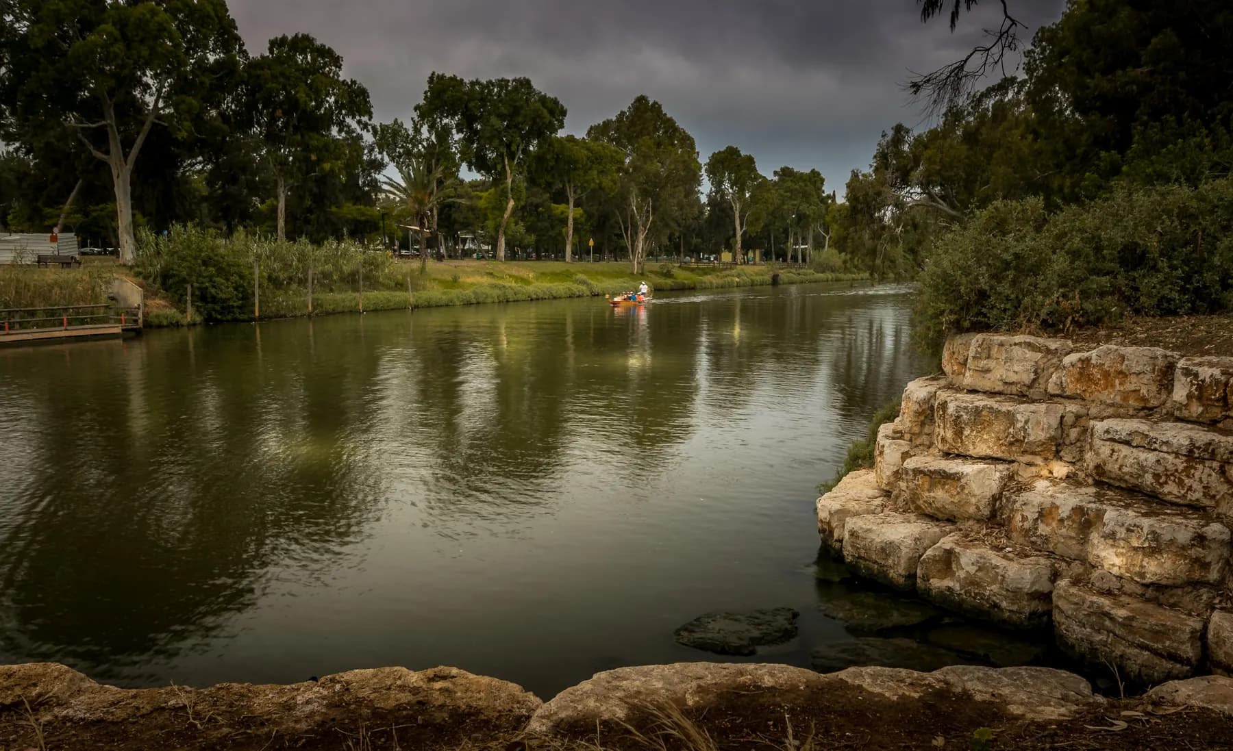 The Yarkon river curving between wooded banks in HaYarkon Park, a small boat out on the water and a stepped stone revetment at the right
