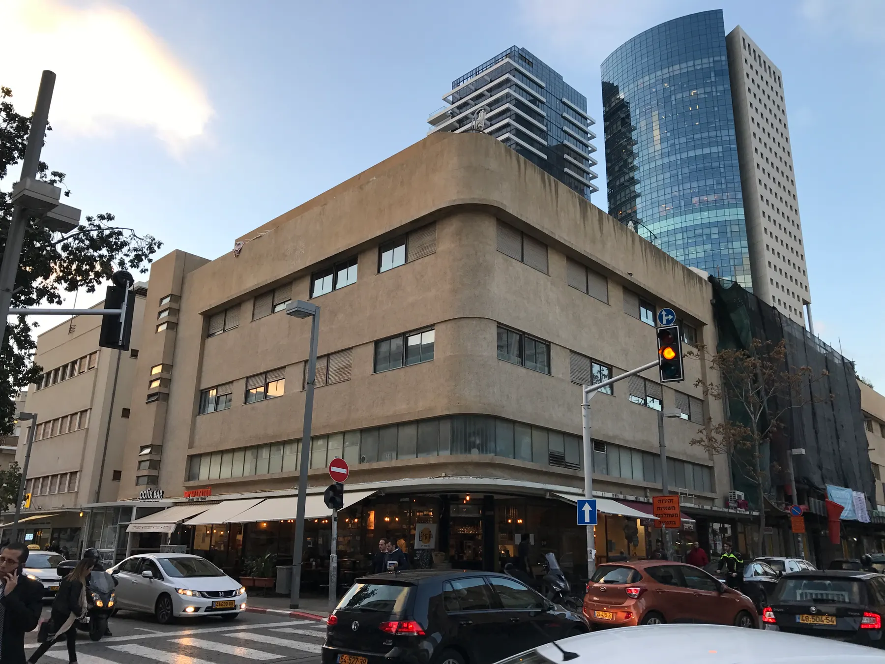 A 1930s International Style corner block in Tel Aviv, its rounded corner wrapped in continuous bands of windows above cafe awnings at street level