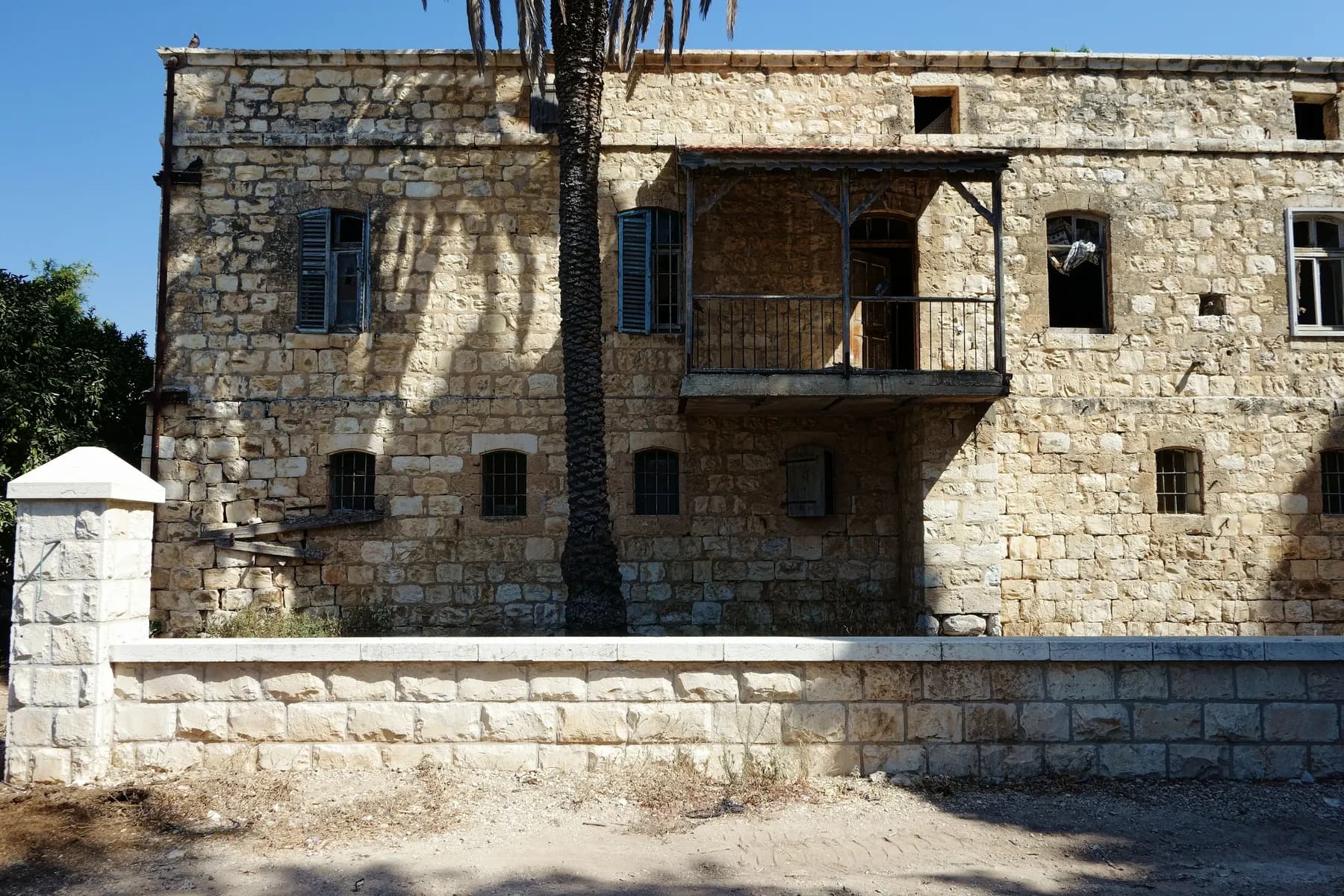 A large two-storey stone house with blue shutters and a covered balcony in Alonei Abba, behind a low stone wall and the trunk of a palm tree.