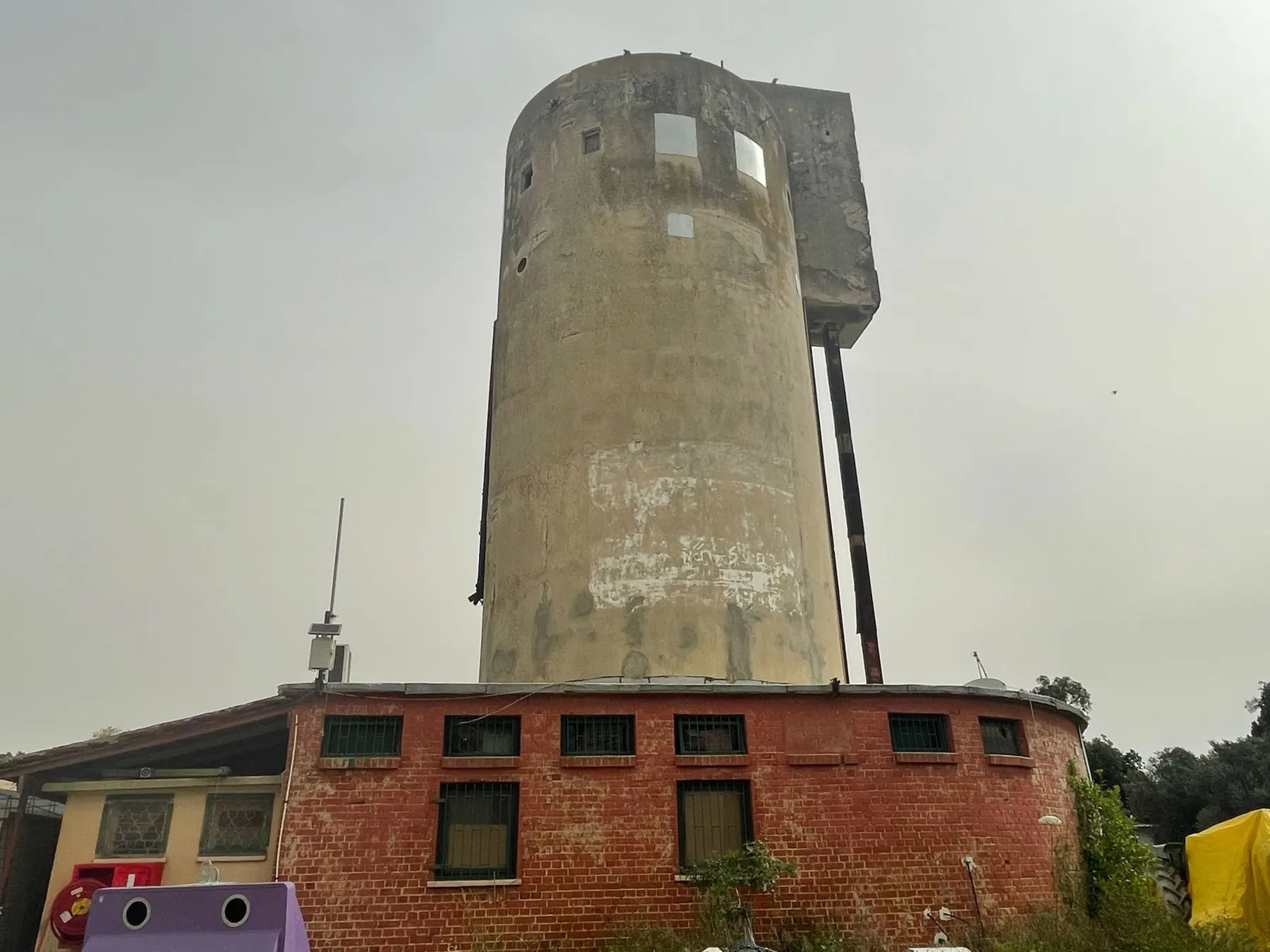The old grain silo of Alonim, a tall round concrete tower rising above a curved red-brick building with barred windows, under a grey sky.