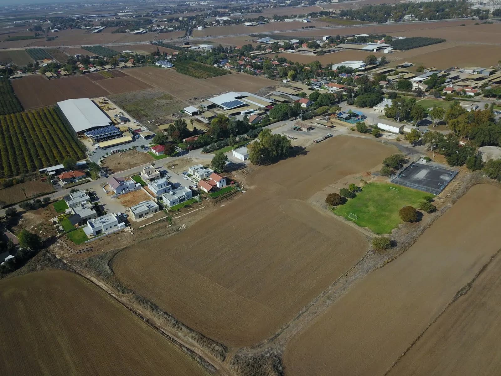 An aerial view of Avigdor, with new white houses and a sports court beside ploughed fields and the older houses and farm buildings among trees beyond.