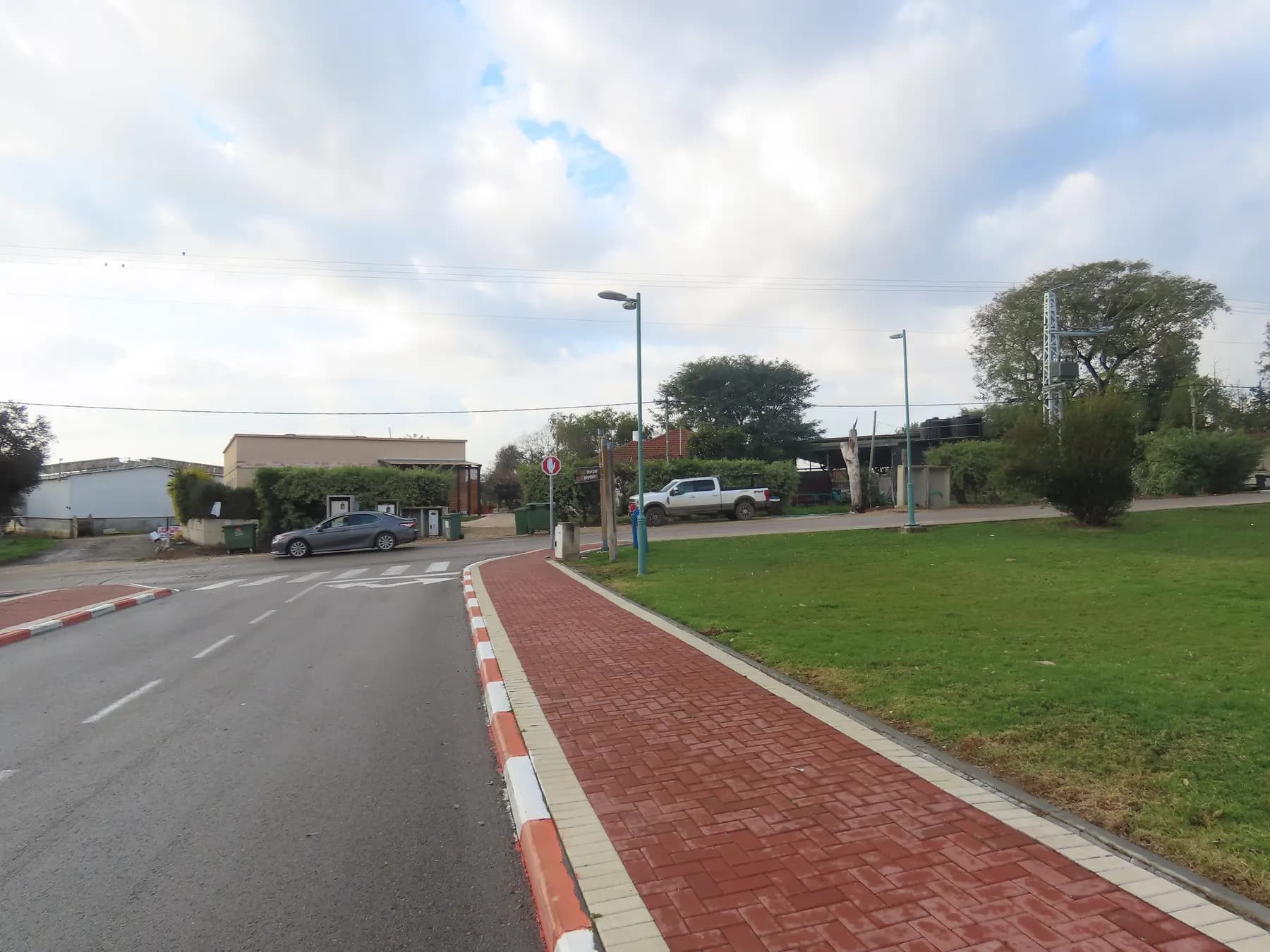 A quiet street in Beit Hilkia with a red brick pavement, a wide lawn and low buildings under a cloudy sky.