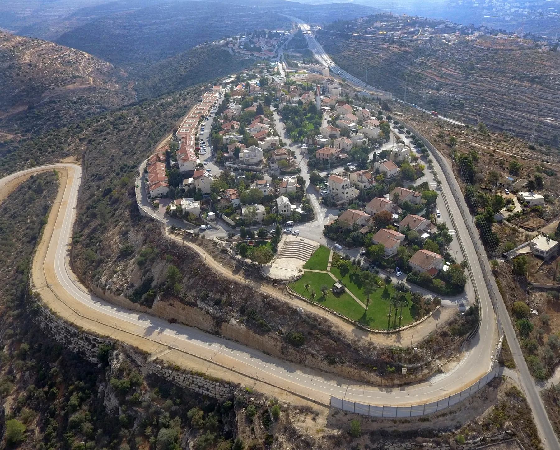An aerial view of Beit Horon, with red-roofed houses, a lawn and an amphitheatre on a hilltop ringed by a winding road and terraced hillsides.